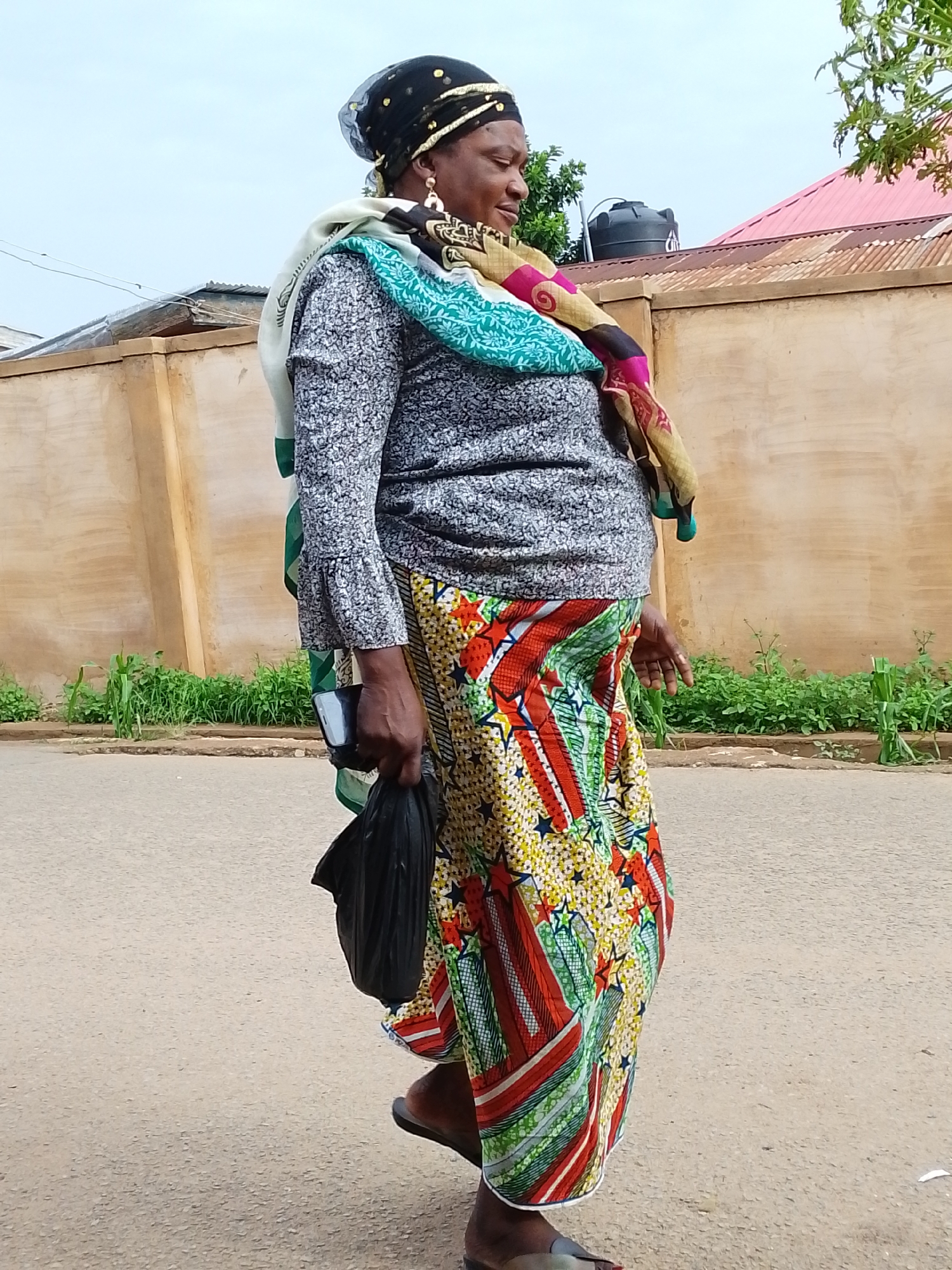 A woman is walking on a street in Jos, Nigeria. She is wearing a gray patterned long-sleeved shirt, a long colorful patterned skirt, a black head covering with gold accents, and a colorful scarf draped around her shoulders. She is carrying a black plastic bag in her left hand and wearing sandals. The street is paved, and there is a wall on the right side of the image with some green plants growing along the base. A water tank is visible on top of the building behind the wall. The sky is overcast. A house with a red roof is also visible.