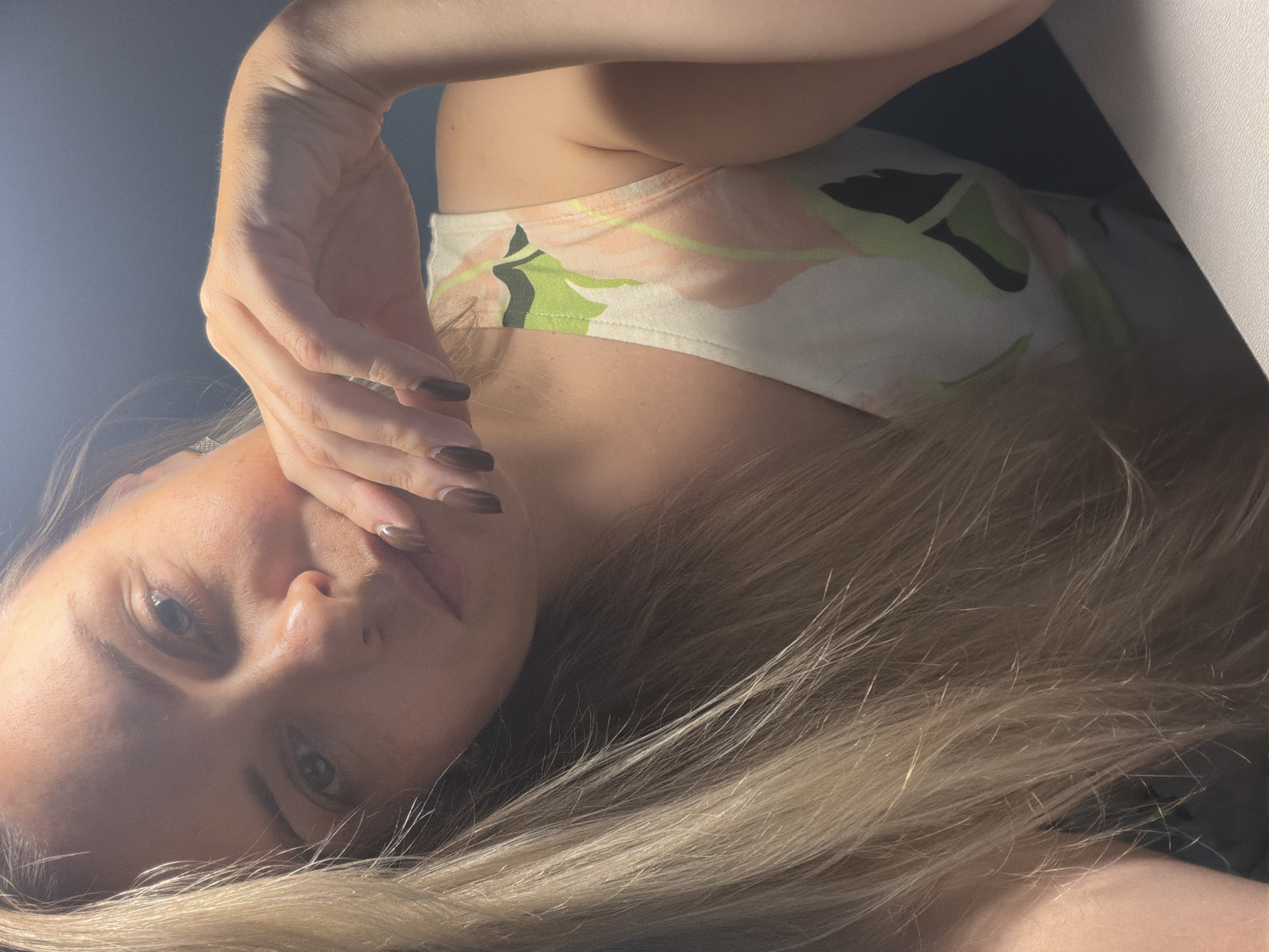 This is a close-up, vertically oriented, low-angle photograph of a young woman lying down. Her face is partially obscured by her right hand, which is raised to her forehead. She has fair skin, light-colored eyes, and long, blonde hair that is spread out around her. She is wearing a tank top with a floral pattern featuring pink, green, and black elements on a white background. The lighting suggests it might be daytime, possibly indoors with artificial light creating a dramatic effect. The overall mood appears contemplative or perhaps tired, given the pose and facial expression. There is no visible text in the image. The location context is Votkinsk, Russia, however, there are no explicit visual cues in the image to confirm this location.