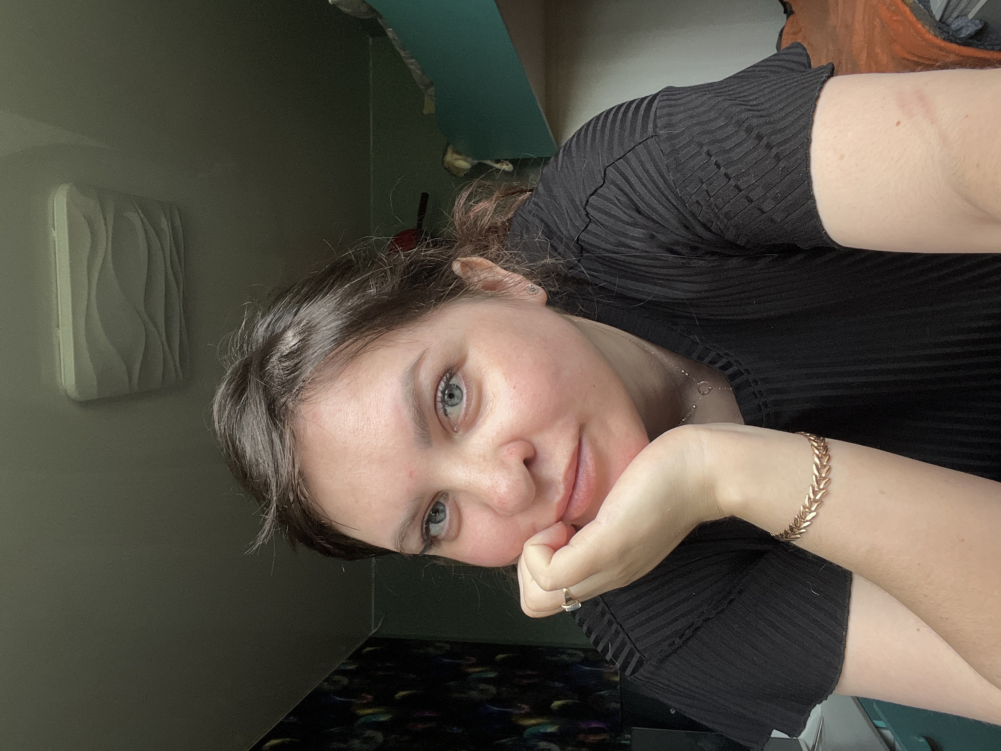 A woman with fair skin, brown hair, and light-colored eyes is posing for a selfie. She is wearing a black, ribbed top and a gold bracelet on her right wrist. Her left hand is resting under her chin, and she is wearing a ring on her left ring finger. The background includes a gray wall with a white, wavy textured decoration, a teal-colored shelf, and other objects that are out of focus. The location is not provided in the image.