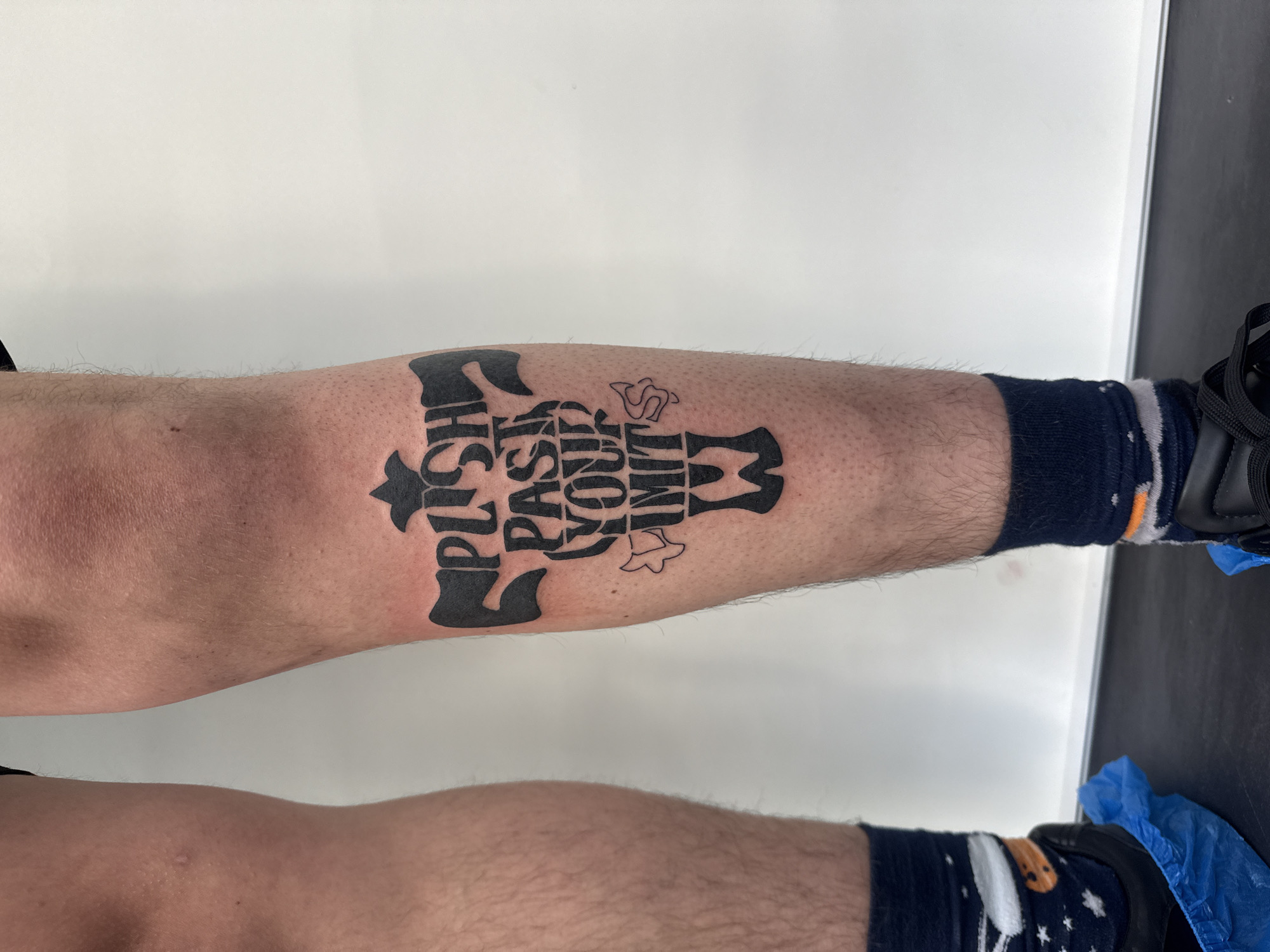 This image presents a close-up, vertical view of a person's lower legs and feet, centered on a new or freshly done tattoo. The setting is indoors, characterized by a bright, clean white background, suggesting a sterile environment like a tattoo studio or medical office.
The main subject is a large, black, text-based tattoo prominently displayed on the front of the person's right lower leg. The tattoo features highly stylized, bold, and wavy lettering that forms a unified, somewhat organic shape. The text reads "PUSH PAST YOUR LIMIT". The word "PUSH" is arranged vertically on the left side of the design, while "PAST YOUR LIMIT" is stacked vertically to its right, with "PAST" at the top, "YOUR" in the middle, and "LIMIT" at the bottom. Several smaller, uncolored outlines, including a stylized "S" and a bell-like figure, are integrated into the overall black ink design. The skin directly around the tattoo appears recently shaven, highlighting the crispness of the black ink against the lighter skin.
The person's legs show visible body hair outside the tattooed area. Both feet are adorned with dark blue socks; the sock on the left leg displays a pattern of white stars and what appears to be a cartoon rocket or moon. The right foot wears a dark shoe, likely a sneaker, which is covered by a blue plastic shoe cover, commonly used for hygiene or protection. The lighting is bright and even, suggesting an indoor, well-lit space. The activity depicted is likely the immediate aftermath of a tattoo session. The location of this event is Sofia, Bulgaria.