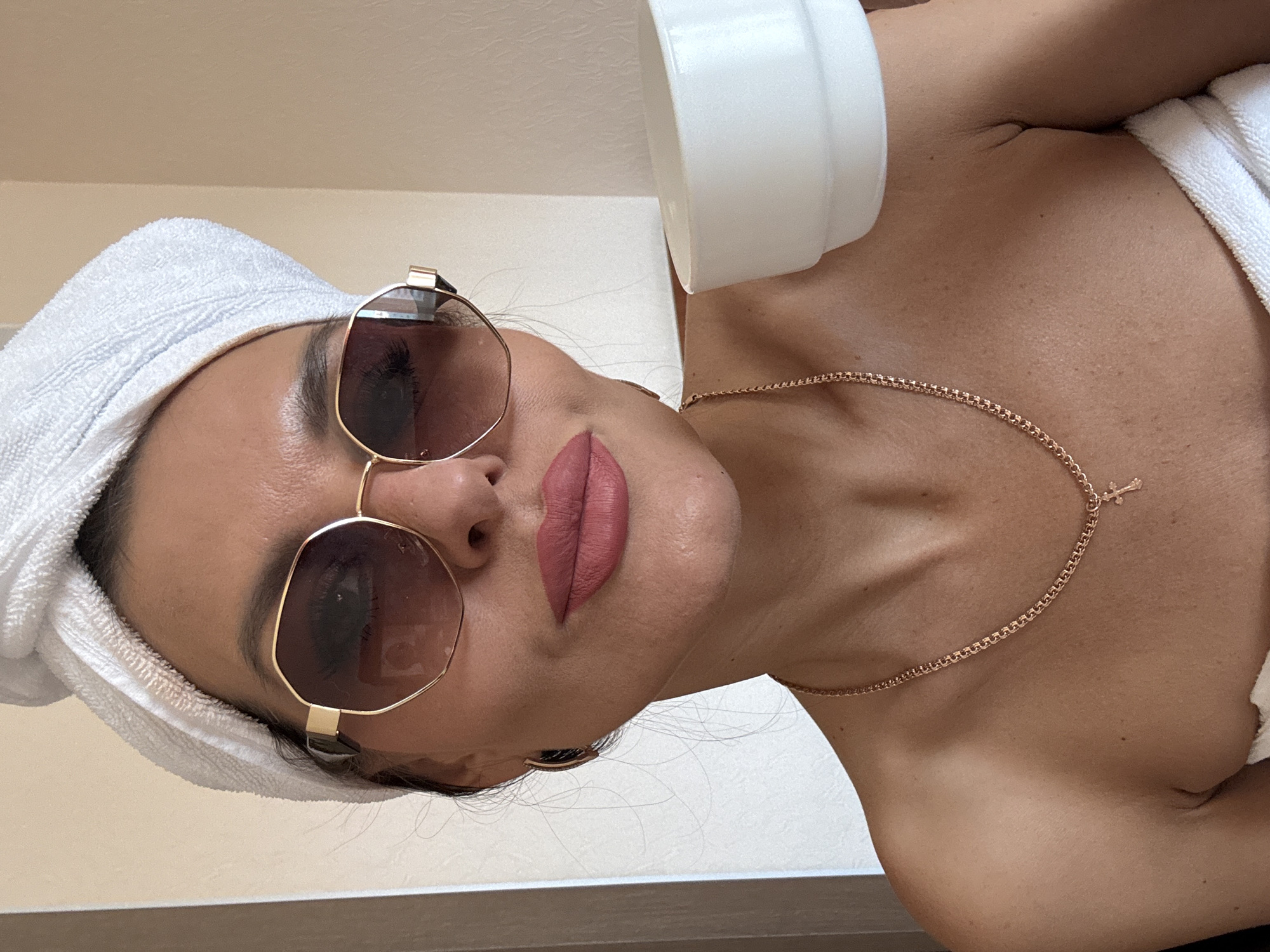 A woman with sunglasses and a towel wrapped around her head is lying down. She is wearing a gold necklace with a small cross pendant. A white cylindrical container is visible in the upper part of the image. The setting appears to be indoors, possibly a spa or salon. There is no information available to discern the location as Perm, Russia.