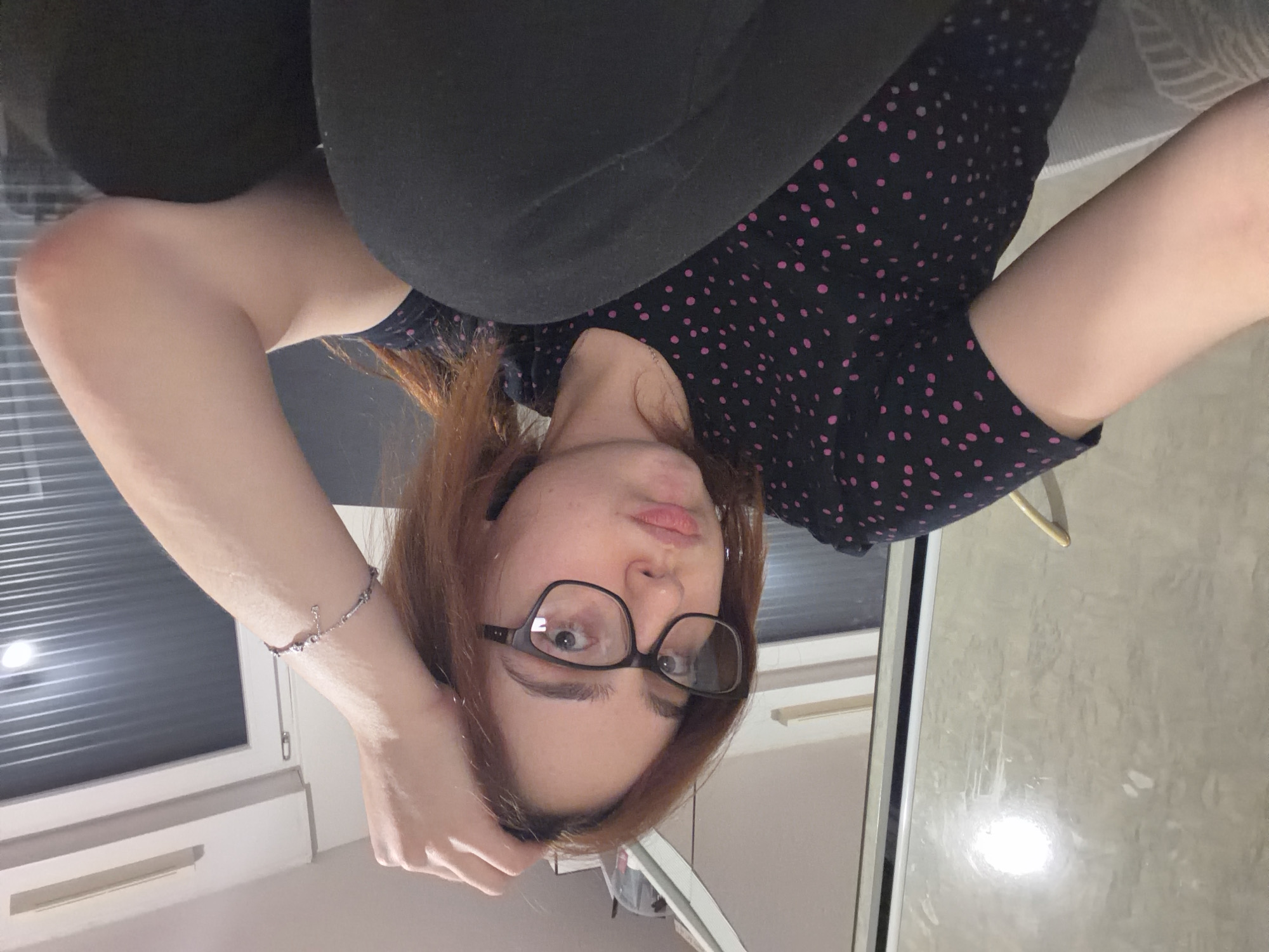 A woman with auburn hair and glasses is angled in the shot, wearing a black shirt with pink polka dots and black leggings. Her left hand, adorned with a bracelet, rests near her forehead. In the background, a window with blinds is visible. The scene appears to be indoors, likely in a residential setting. It is not possible to determine the location within Strasbourg, France, from this image alone.