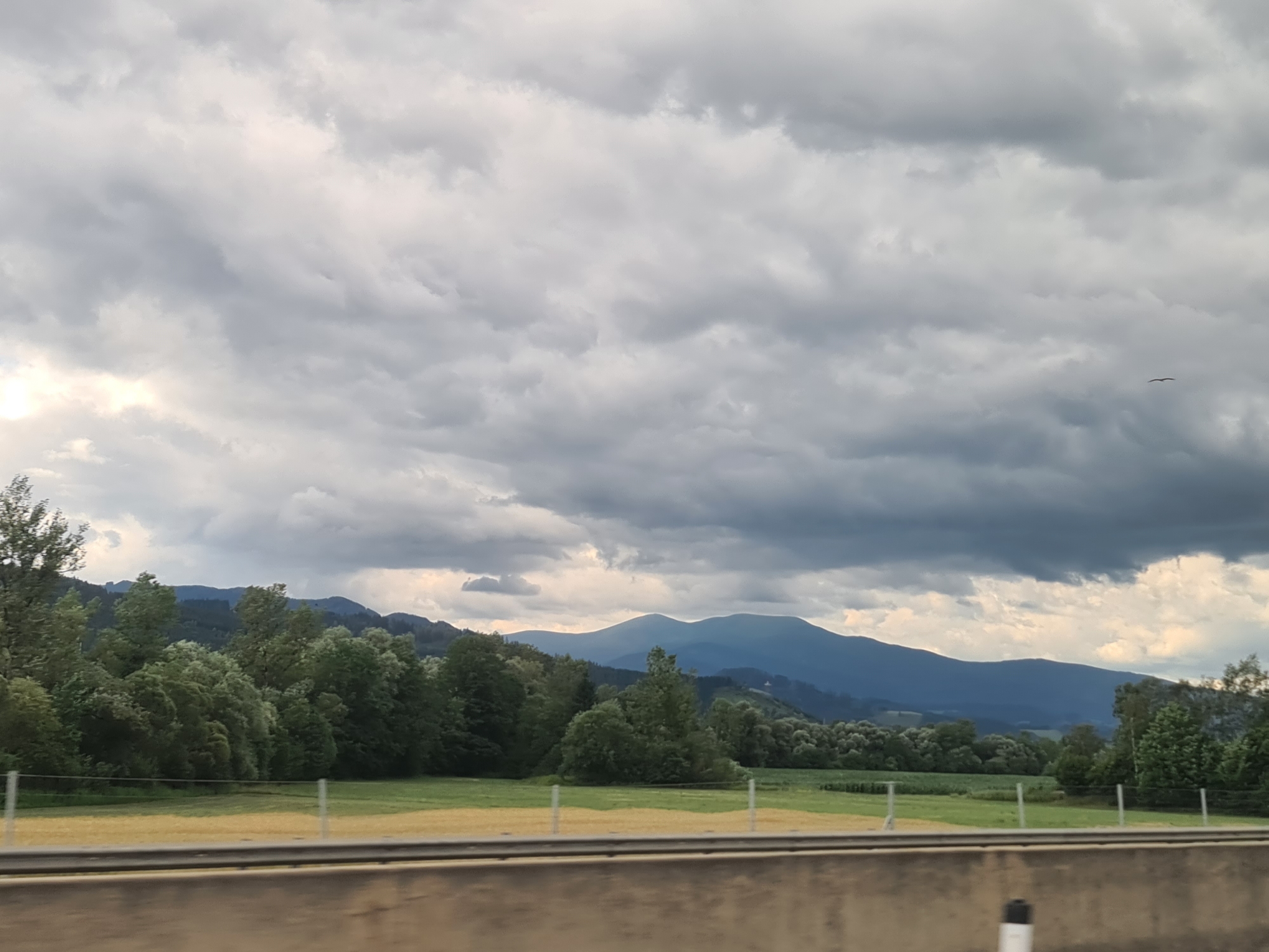 The image depicts a cloudy sky over a landscape of rolling hills and mountains. In the foreground, there is a blurred concrete barrier, suggesting the photo was taken from a vehicle. Beyond the barrier, a grassy field with some yellowing vegetation is visible, bordered by a fence. A dense line of trees forms a mid-ground, with varying shades of green indicating different types of foliage. In the background, layered blue mountains recede into the distance under a dramatic sky filled with heavy grey and white clouds. A single bird is visible in the upper right quadrant of the sky. The scene is devoid of people and specific identifiable structures, making it a general landscape view. The location is identified as Kobenz, Austria, a municipality in the district of Murtal in Styria.
