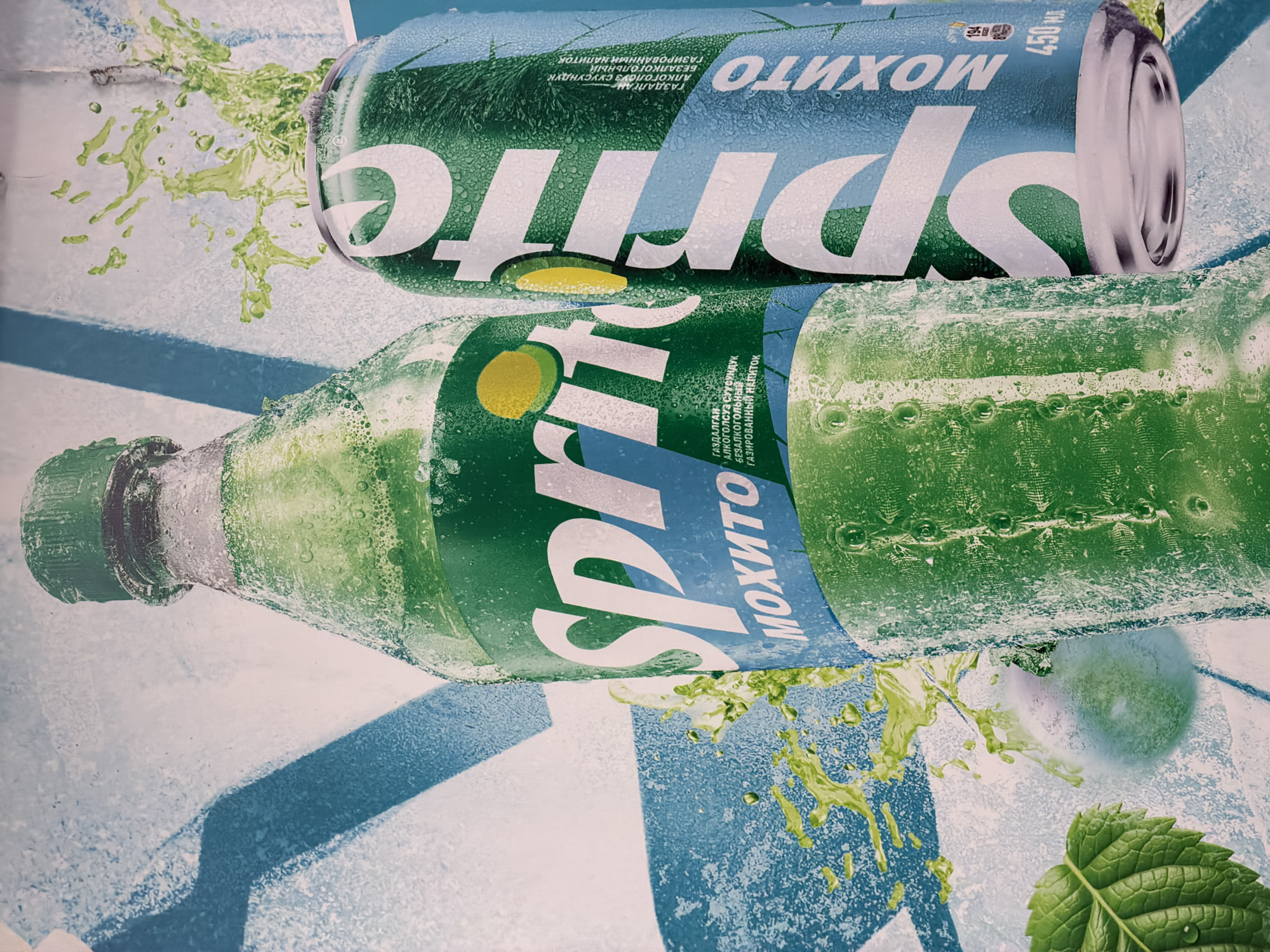 This detailed image, captured in Unknown, Unknown, displays a promotional advertisement for Sprite Mojito. The foreground features a large, green plastic Sprite bottle on the left and a slender, green aluminum Sprite can on the right, both depicted with visible condensation and water droplets, indicating a cold temperature.
The bottle has a green screw cap and a primary white "Sprite" logo, which includes a yellow lemon/lime slice icon. Below the logo, a blue label segment reads "МОХИТО" (Mojito) in white Cyrillic text.
The can also features a prominent white "Sprite" logo oriented vertically, with a blue label segment displaying "МОХИТО" (Mojito) in white Cyrillic text. Additional Cyrillic text, including "ГАЗАЛҒАН," "АЛКОГОЛЬСУЗ СУСУНДУК," and "БЕЗАЛКОГОЛЬНЫЙ ГАЗИРОВАННЫЙ НАПИТОК" (Non-alcoholic carbonated drink), is visible, along with "450 МЛ" (450 ML). The top of the can shows a small amount of green liquid splashing outwards.
The background consists of a textured, light-colored surface with broad, frosty blue stripes running diagonally. Dynamic splashes of a green liquid are visible around both the bottle and the can, contributing to a sense of refreshment. In the lower-left corner, a single, vibrant green mint leaf with visible veins and water droplets is present. A subtle, blurred green circular object, possibly a lime or mint element, is discernible towards the bottom center of the frame. The perspective is slightly angled, looking down upon the beverages.
