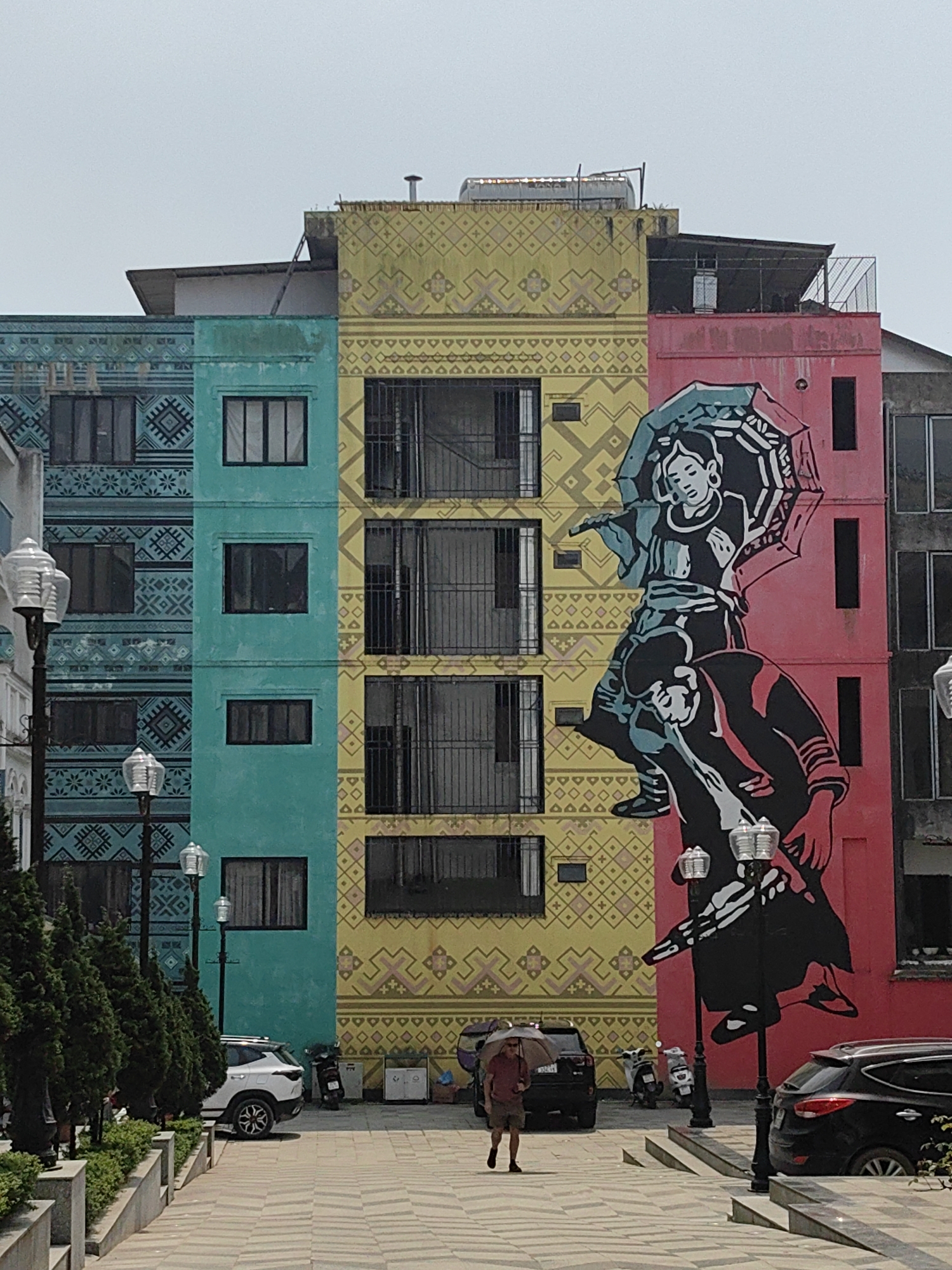 The image displays a street scene in Sa Pa, Vietnam, centered on a row of multi-story buildings. From left to right, three main buildings are visible with facades painted distinctively: the leftmost is teal with dark geometric patterns, the central building is yellow with lighter geometric patterns, and the rightmost is solid pink. A partial view of a fourth, grey building is present on the far right. All buildings feature multiple rectangular windows with dark frames, and those on the yellow building have black metal security grates. The rooftops exhibit various utility structures, including vents, a solar water heater, and metal railings.
A large mural spans the yellow and pink building facades, depicting two stylized figures in traditional attire. The upper figure, smaller in stature, holds an open teal and white umbrella. This figure is positioned atop the shoulders or back of the lower, larger figure. The lower figure is shown from the waist up, supporting the upper figure. The mural is rendered predominantly in black and white, with teal elements on the umbrella and the upper figure's clothing, and subtle red accents.
In the foreground, a patterned tiled plaza extends from the bottom of the frame towards the buildings. A landscaped median runs along the left side of the plaza, featuring multiple black streetlights with lantern-style heads and planted small evergreen trees and shrubs. Several vehicles are parked along the side, including a white SUV, a black SUV, and a scooter. A single individual, dressed in a red short-sleeved shirt and shorts, walks across the tiled plaza from left to right, holding an open beige umbrella for sun protection. The sky is bright and clear.