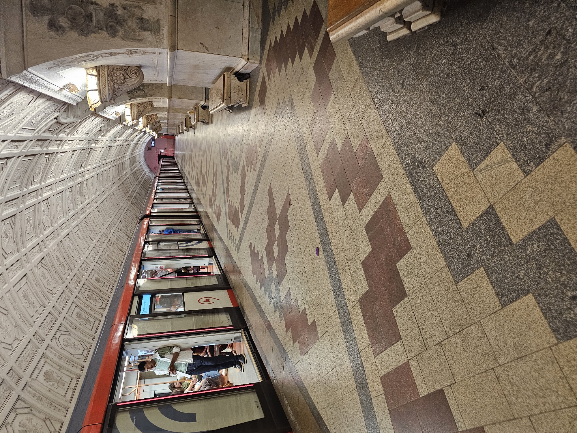 This image depicts the interior of a Moscow Metro station, specifically the Prospekt Vernadskogo station on the Sokolnicheskaya Line. The perspective is from the platform, looking down the length of the curved tunnel. A train with open doors is stationary on the tracks to the left, with several passengers visible both inside and on the platform.
The station's architecture is characterized by a high, ornate ceiling with a repeating patterned design. The platform is paved with a mosaic of beige, dark gray, and maroon tiles arranged in a geometric pattern. To the right of the platform, there are arched alcoves with decorative elements and wall sconces providing illumination. Stone benches, also ornately carved, are placed along the wall in front of these alcoves.
The lighting is a mix of ambient station lights and the illuminated train interior. The overall impression is one of grand and classical underground architecture.