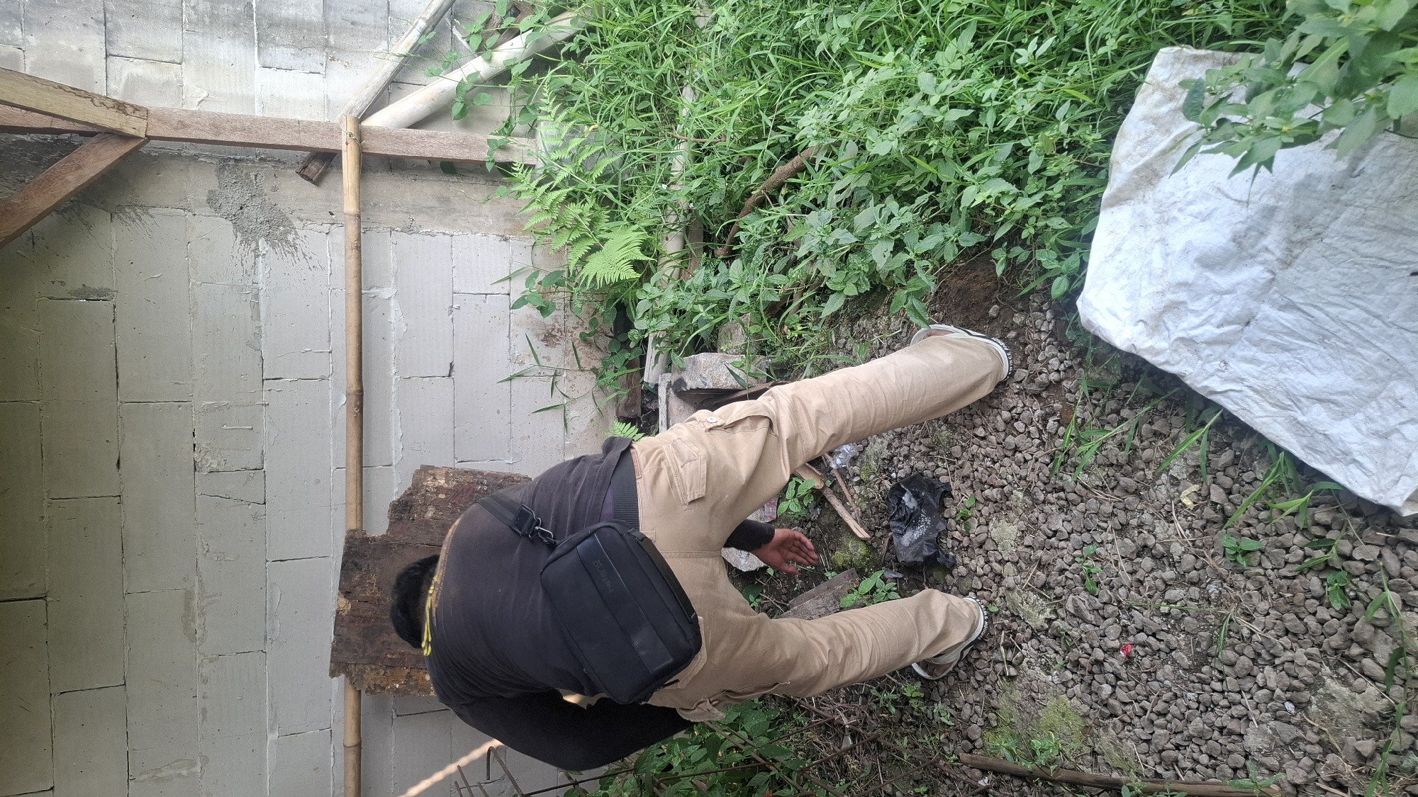 A person is bending over in a yard outside of a building. The building has a cinder block wall and a wooden roof structure. The ground is covered in gravel and grass. There is a black bag on the ground and a white tarp in the lower right corner. The person is wearing a black long-sleeved shirt and tan cargo pants. They have a black bag slung across their chest. The person is reaching for the black bag on the ground. The scene is located in Rahong, Indonesia.