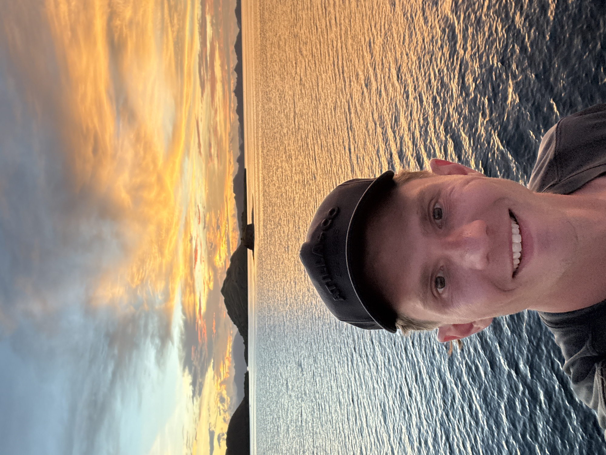 A selfie shows a smiling man wearing a baseball cap on a body of water during sunset. The water is reflective with orange and blue colors. There is a rocky landmass on the left side of the image with a vibrant orange sunset sky. Location: Unknown, Indonesia.