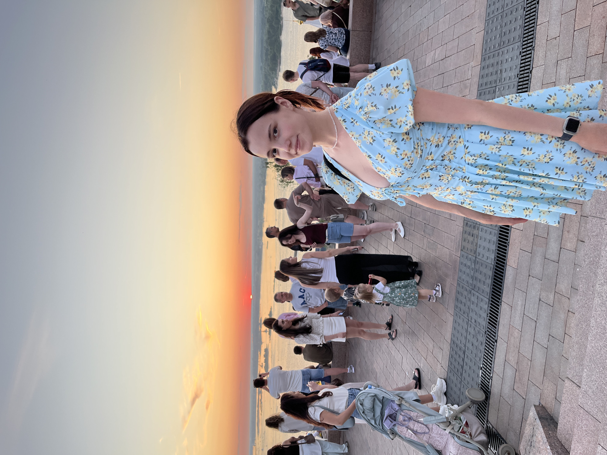 A woman wearing a light blue floral dress stands in the foreground, facing the camera with a slight smile. She has dark hair pulled back and is wearing a necklace. Behind her, a crowd of people is gathered on a paved area overlooking a wide river. The sun is setting over the water, casting a warm orange glow across the sky. In the background, there are trees and buildings. The overall scene appears to be a public gathering spot, likely a scenic viewpoint in Nizhny Novgorod, Russia, given the presence of the Kremlin or other historical architecture visible in the distance.