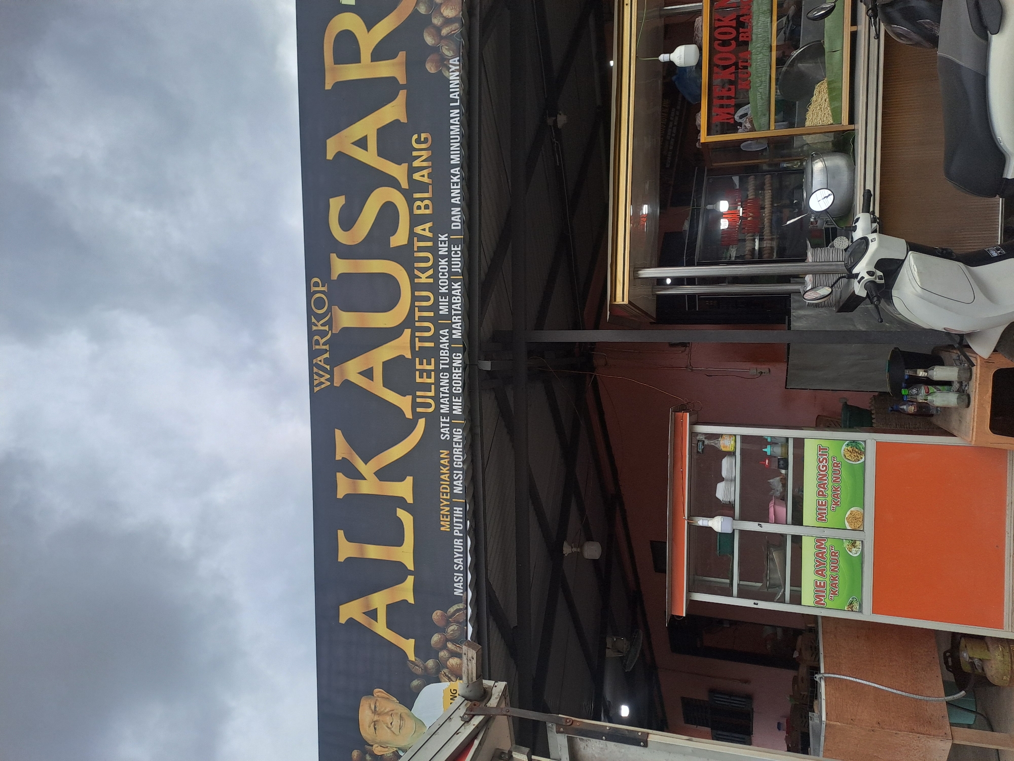 Here is a description of the image:
The image shows a street scene, likely in Tingkeummanyang, Indonesia, given the writing on the signs. A large sign reading "WARKOP ALKAUSAR" dominates the left side. Below this is "ULEE TUTU KUTA BLANG." Below the sign are listed various food items such as "SATE MATANG TUBAKA," "NASI SAYUR PUTIH," "NASI GORENG," and "JUICE." A man's image is present at the bottom of the sign.
On the right side of the photo, a scooter is parked. There's a food stall with an orange exterior, displaying "MIE AYAM" and "MIE PANGSIT" offerings. A portion of a window is seen above the scooter with the text "MIE KOCOK." The sky appears cloudy.