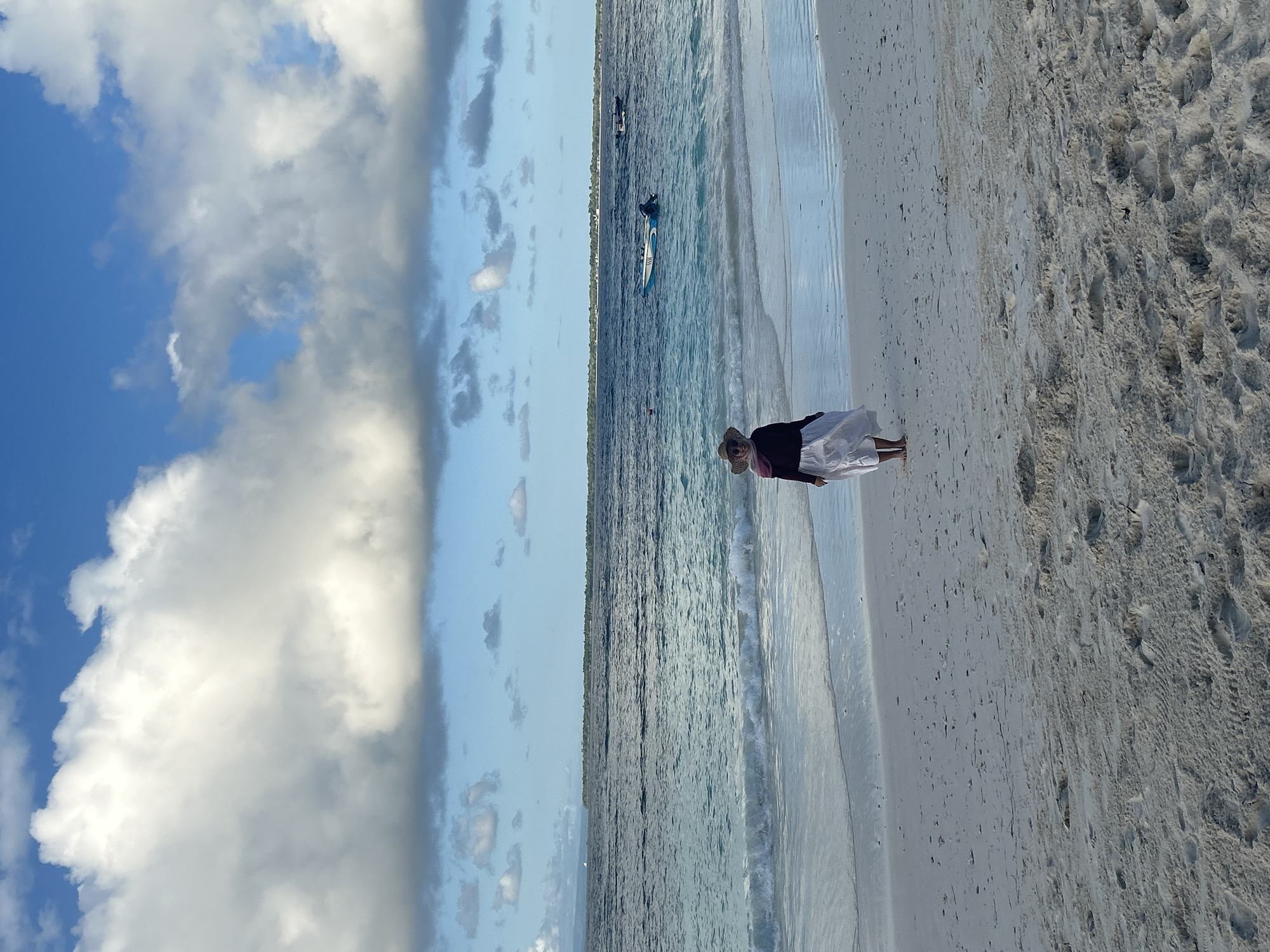 A woman wearing a straw hat, dark top, and white skirt stands on a sandy beach facing the ocean in A R A, Indonesia. The ocean water is a turquoise blue with gentle waves. In the distance, several boats are visible on the water, and a long strip of land is on the horizon. The sky is blue with scattered white and gray clouds. The sand is light in color and shows imprints of footprints.