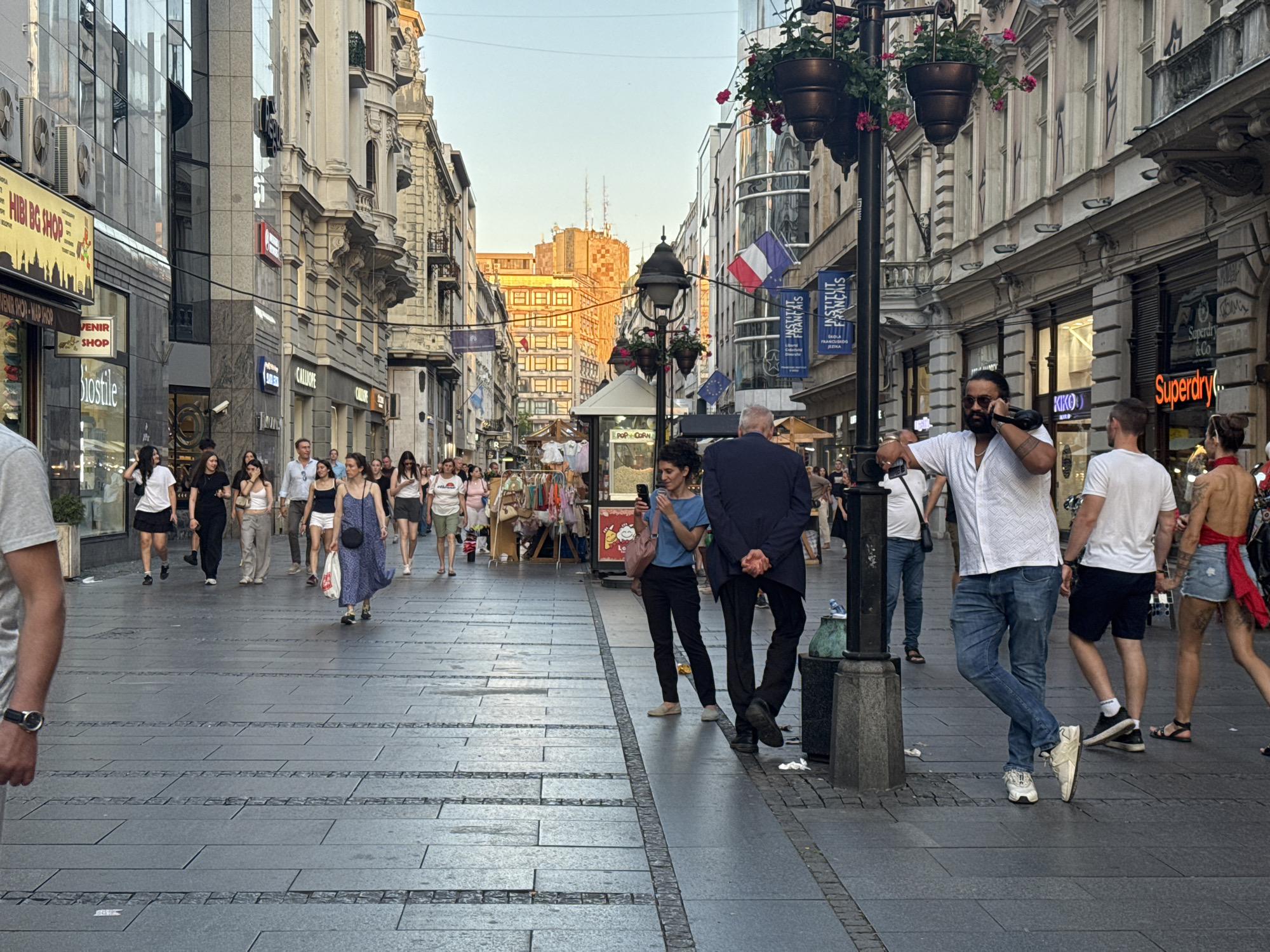 The image depicts a bustling pedestrian street scene in Belgrade, Serbia, likely Kalemegdan Street given the architectural style and ambiance. The sun is setting, casting a warm glow on the buildings and the crowd.
On the left side of the image, there are shops with glass facades displaying their merchandise. Several individuals are walking in the same direction, some browsing shop windows. A person on the far left is partially visible, wearing a grey t-shirt and a watch.
The center of the image features a wide pedestrian walkway paved with dark grey stone tiles. More people are seen walking and interacting. A woman in a blue dress is walking towards the viewer, carrying a black crossbody bag. In the background, there are more shops and buildings with ornate facades.
On the right side, a group of people are also walking. A man in a white shirt and blue jeans is standing and talking on a mobile phone, with his body leaning slightly against a lamppost. Another man in a white t-shirt and dark shorts is walking away from the viewer. A woman in a red top and denim shorts is also walking away. Lampposts decorated with hanging flower baskets are placed along the street. Banners with text and flags are visible, suggesting cultural or commercial establishments.
The background shows buildings with a mix of modern and classical architecture, some with balconies and intricate detailing. The sky is a clear blue with a hint of orange from the setting sun. The overall scene conveys a lively urban atmosphere, with people enjoying their evening on a popular city street.