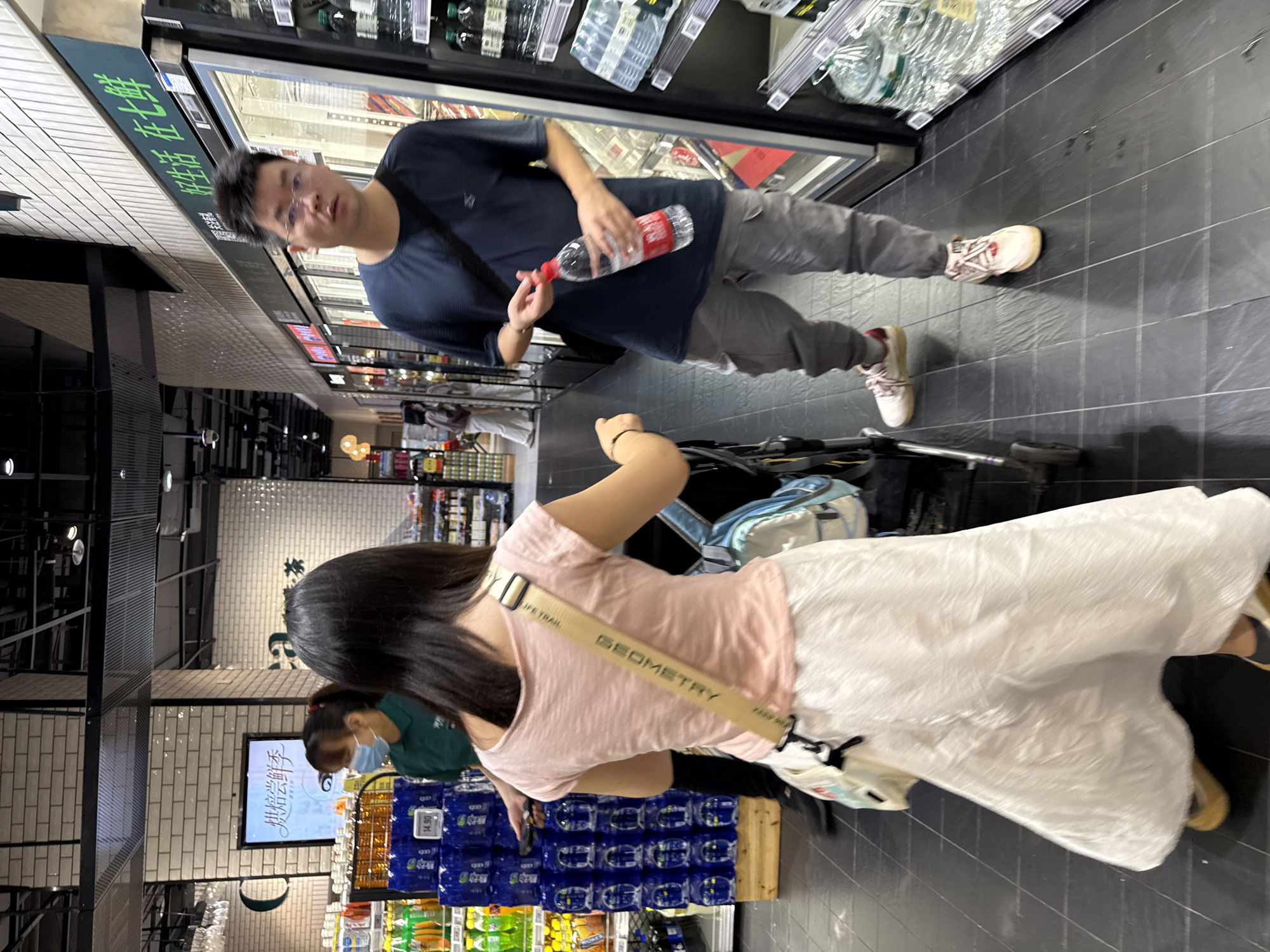 The image depicts an indoor scene, likely a supermarket, with a black tiled floor. In the foreground, a woman wearing a pink t-shirt and a flowing white skirt is seen from behind, pushing a baby stroller. A beige strap with the word "GEOMETRY" is slung across her chest. To her right, a man in a dark blue t-shirt and grey cargo pants is holding a clear plastic bottle of water. He is wearing glasses and white sneakers. In the background, a woman wearing a green shirt and a face mask is standing near a display of bottled drinks. A sign above a refrigerated display case reads "好生活 在七鲜", which translates to "Good life at Qi Xian". This suggests the location is likely a "Qi Xian" supermarket in Haizhu District, China. The environment appears well-lit with fluorescent lighting from above.