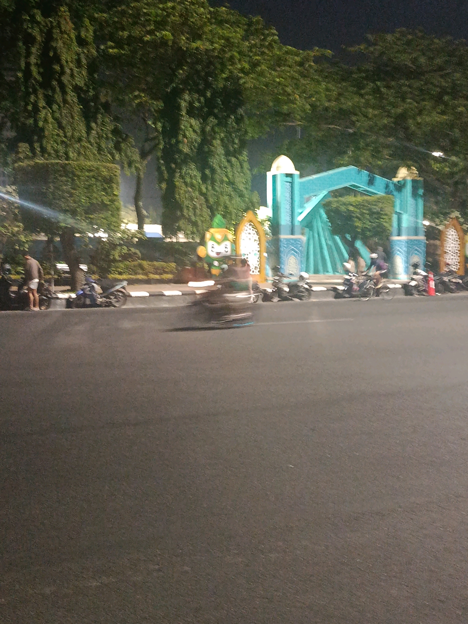 This nighttime photograph captures a street scene in Semarang, Indonesia, near the Simpang Lima monument. The image features a turquoise archway, possibly part of a decorative entrance or monument, with ornate gold and white embellishments on either side. In front of the archway, a cartoonish green and yellow mascot character stands. The street is lined with parked motorcycles. A person in a light-colored shirt and shorts is standing next to a motorcycle on the left side of the image. Another person on a motorcycle is blurred in motion in the center foreground, suggesting the photograph was taken from a moving vehicle. The background is dominated by lush green trees, illuminated by artificial lights, creating a slightly hazy ambiance. The asphalt road is visible in the foreground, with a white and grey curb separating it from the parked vehicles.