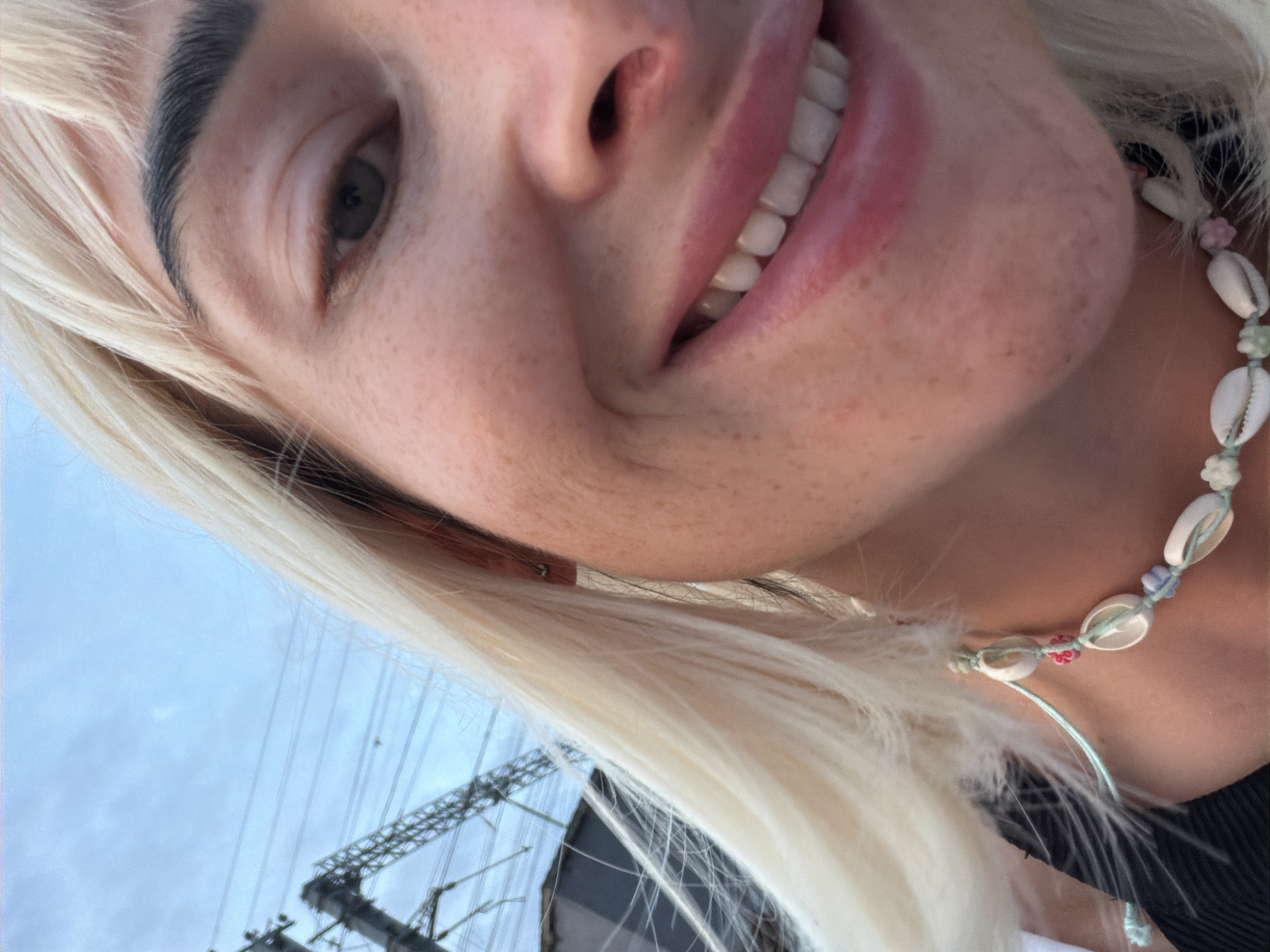 This is a close-up, low-angle selfie of a young woman with blonde hair and fair skin. She is smiling, revealing her teeth. She is wearing a necklace made of shells and beads. The background consists of a pale blue sky with some clouds, and in the distance, there are electrical towers and wires, suggesting an outdoor urban or industrial setting. The lighting is natural, possibly overcast daylight. The image does not provide enough information to determine the specific location in Sochi, Russia.