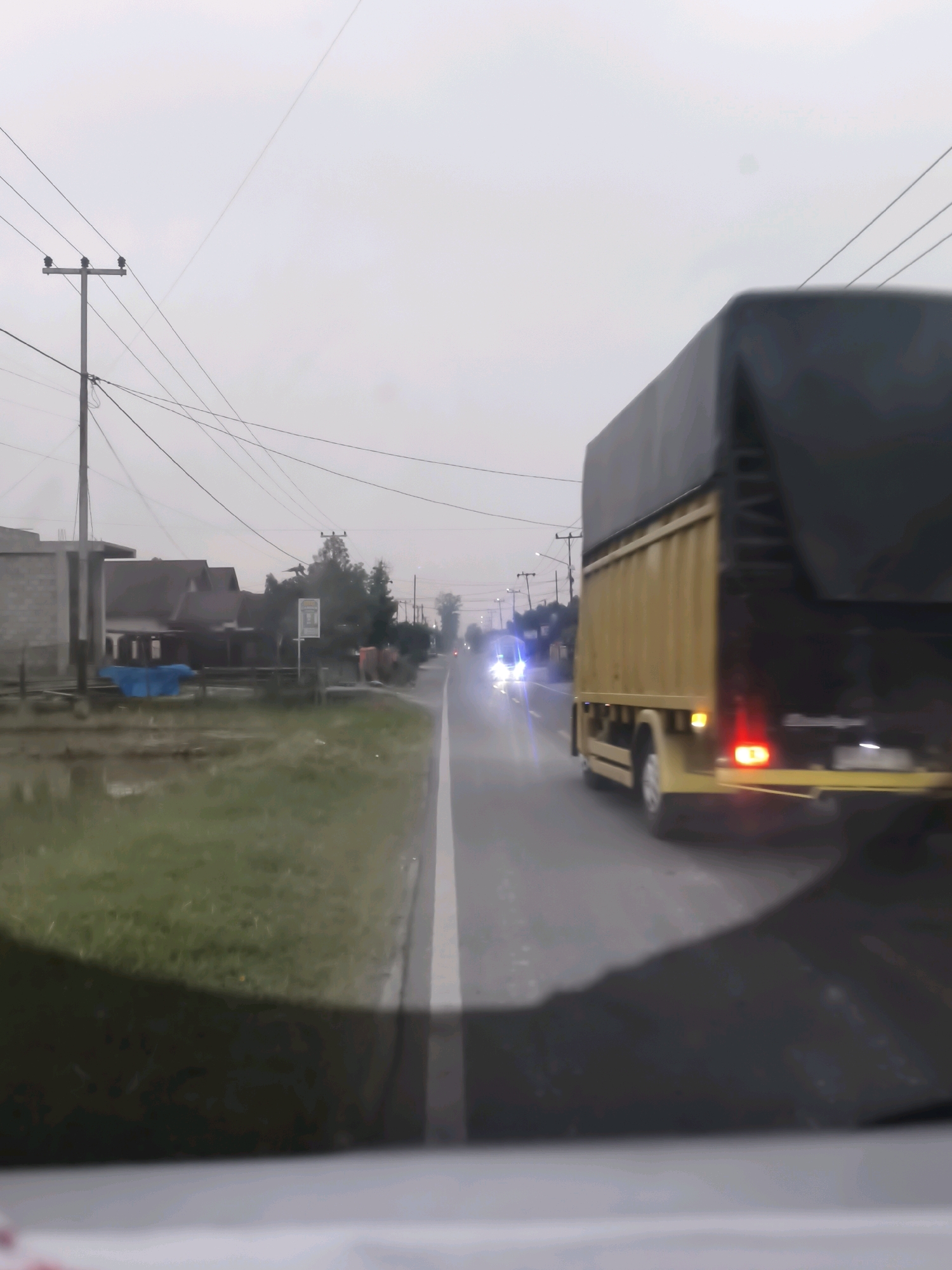 The image depicts a highway scene in Sungai Lebuh, Indonesia, under an overcast sky. The foreground is dominated by the interior of a vehicle, with a dark, shadowy area suggesting the dashboard or hood. A yellow truck with a black tarp-covered cargo area is visible on the right side of the road, moving away from the viewer. Its taillights are illuminated, and a side marker light is also visible. Ahead of the truck, on the left side of the road, are houses and a brick building. Power lines crisscross the sky, supported by utility poles. The road surface appears wet, indicating recent rain or high humidity. In the distance, other vehicles with illuminated headlights are visible on the road. The overall atmosphere is somber and slightly hazy.