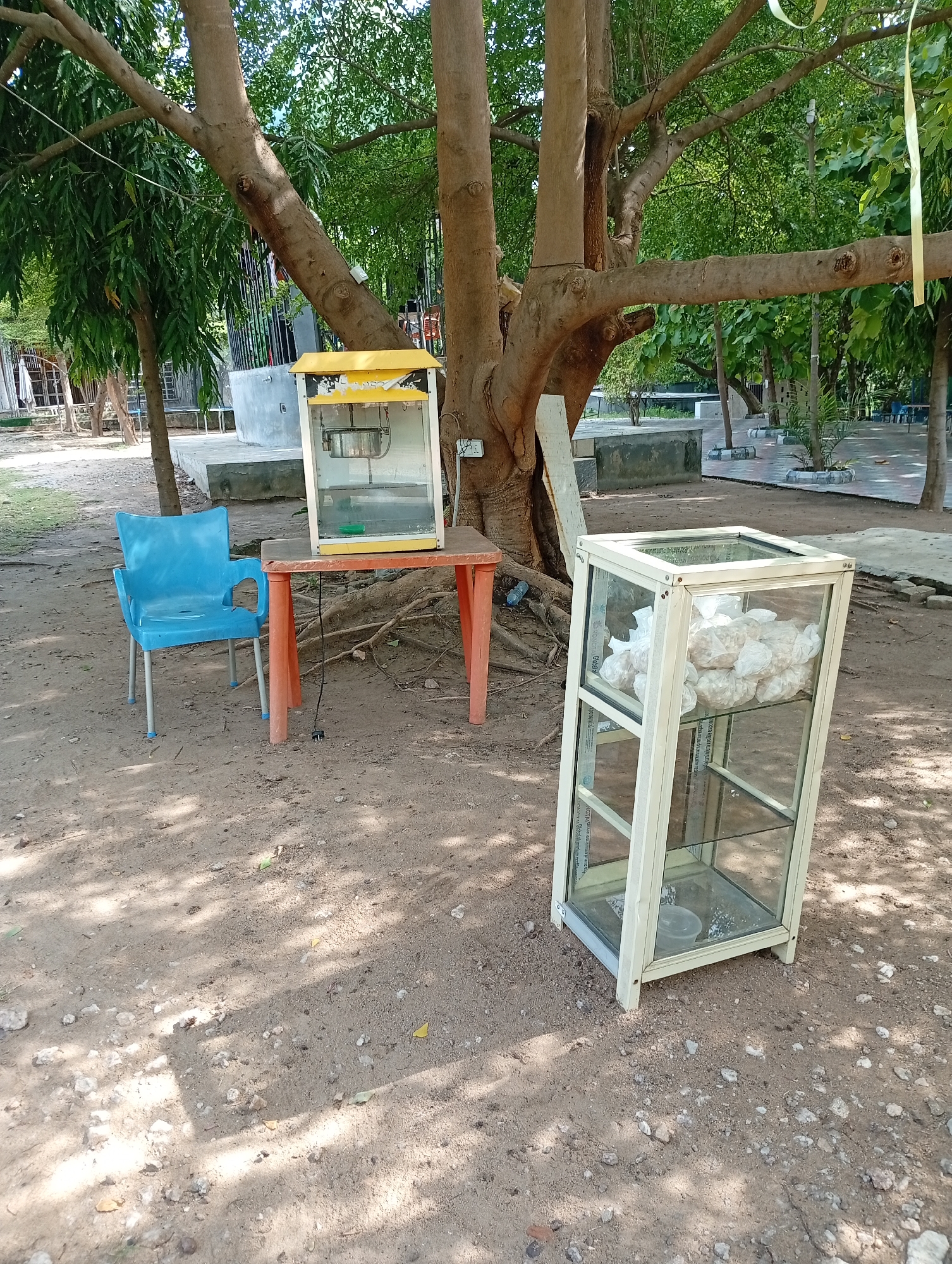 Outside in Adankolo, Nigeria, a popcorn machine sits on an orange table next to a blue plastic chair at the base of a large tree. A glass display case filled with bags of what appear to be snacks stands nearby on the dirt ground. The area appears to be a park or recreational area, with other trees and structures visible in the background.