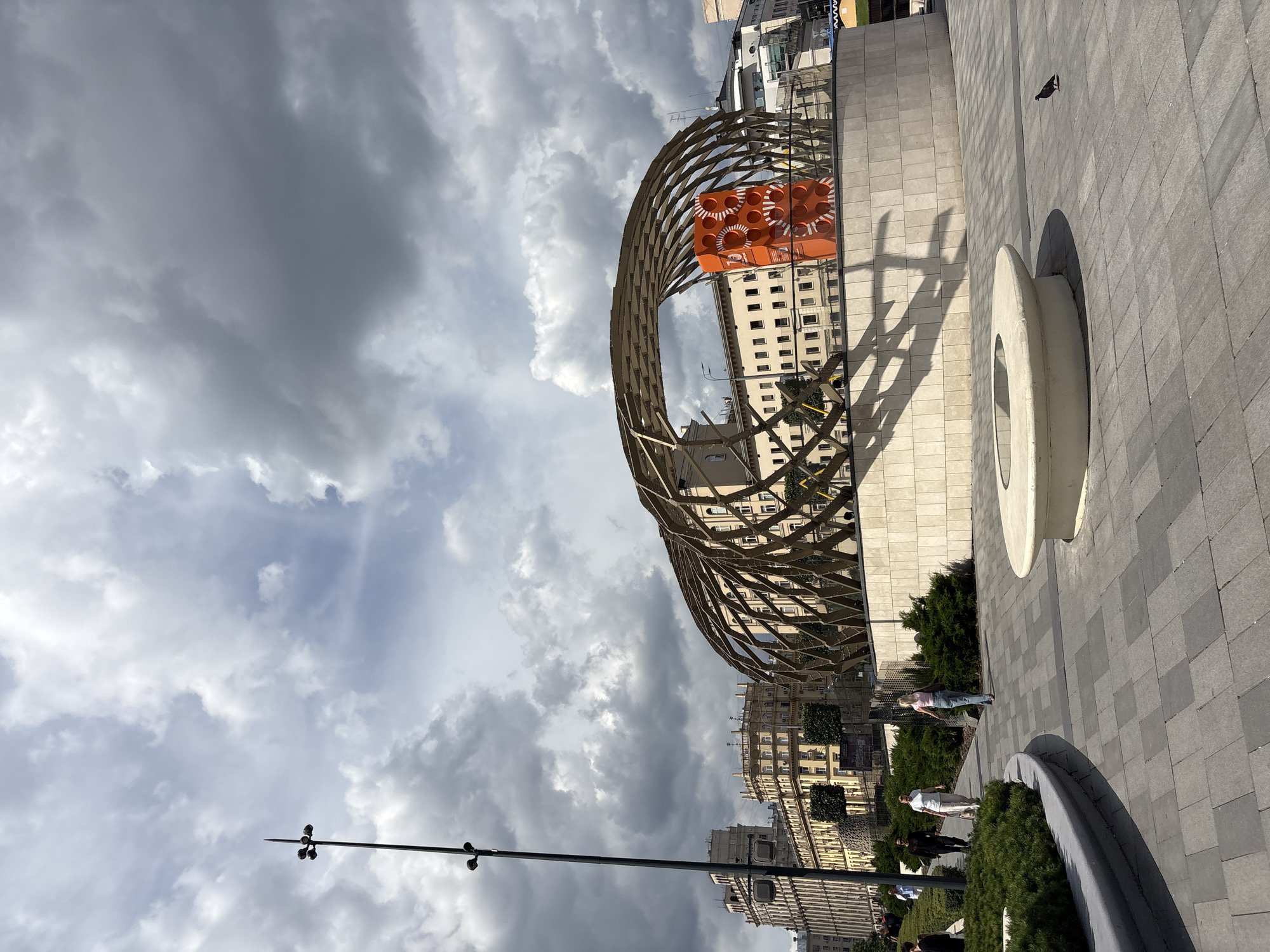 The image shows a public space in Moscow, Russia, likely a plaza or park, with several architectural elements and people. In the foreground, there is a paved area with grey square tiles and a circular concrete structure. To the left, there are manicured green bushes and a curved planter. Several people are walking in the background, dressed in casual attire.
Dominating the midground is a large, modern architectural structure with a wooden lattice roof that curves over a base made of beige stone tiles. Inside this structure, a bright orange, rectangular sculpture with circular cutouts is prominently displayed. Behind this structure, several multi-story buildings with classical architectural features, typical of Moscow, are visible under a dramatic, cloudy sky. A tall, black lamppost with multiple lights stands to the left of the frame. A pigeon is visible in the lower right portion of the image on the tiled ground. The overall scene suggests an urban environment with contemporary art installations and traditional architecture.