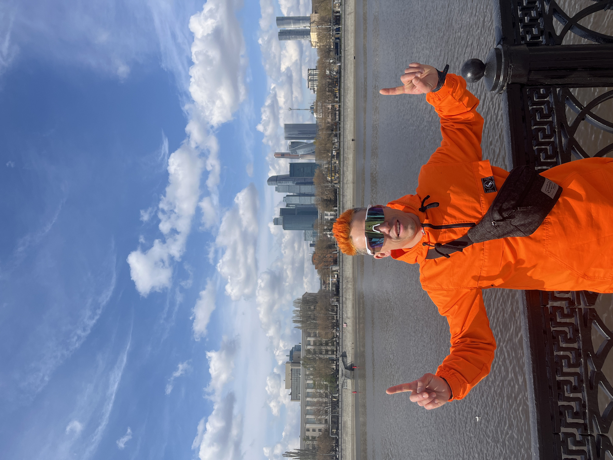 A man with orange hair and reflective sunglasses poses on a bridge in Moscow, Russia, pointing in opposite directions. He is wearing an orange jacket and a dark crossbody bag. Behind him, the cityscape of Moscow is visible with its tall buildings. The sky is blue with white clouds. The man stands next to a black metal railing along the river.