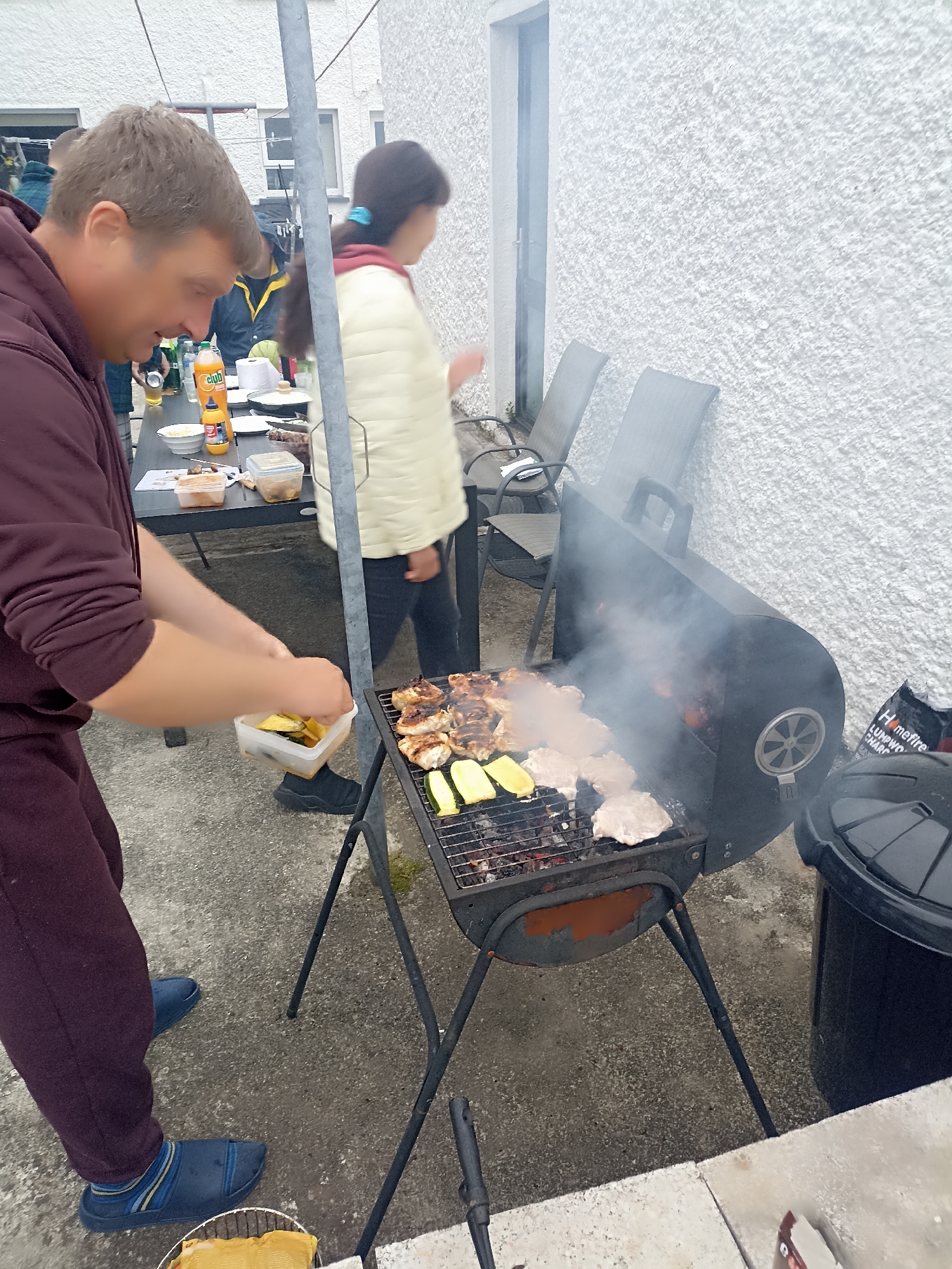 The image captures a casual outdoor gathering in what appears to be a residential area, possibly in Annagry, Ireland, given the context of the query. A man in a maroon hoodie and sweatpants is in the foreground, facing a grill laden with various items, including chicken and zucchini. He is holding a small white container filled with what looks like prepped vegetables or fruit. To his right, a black charcoal grill is actively smoking, with food cooking on its grates. In the background, a woman in a light-colored jacket stands near a table set up with food and drinks, including a bottle of orange soda and a yellow bottle, possibly mustard or sauce. Behind them, a white textured wall of a building and two grey outdoor chairs are visible. The ground is paved with concrete. A large black trash bin is situated to the right of the grill. The overall scene suggests a backyard barbecue or a similar informal outdoor meal.