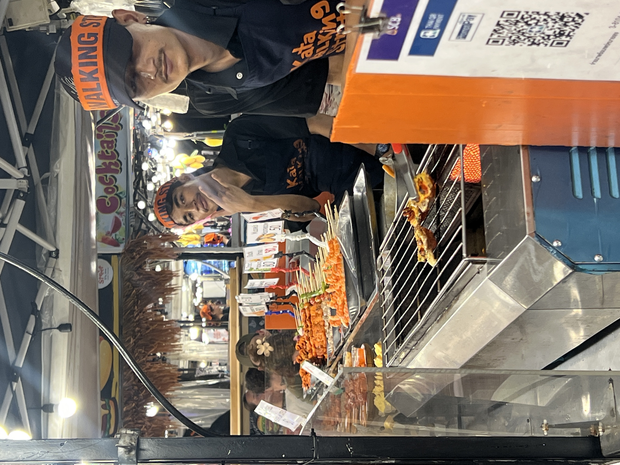 The image depicts a street food stall in Karon, Thailand. Two young men, likely vendors, are visible. One is wearing a dark t-shirt with orange lettering that says "KATA WALKING ST" and a dark cap with an orange band that also reads "WALKING ST". The other person appears to be wearing a similar uniform. They are standing behind a display of various food items on skewers, which are being grilled. The food appears to be meat, likely chicken or pork, and is arranged on a metal grill.
The stall is set up outdoors, suggested by the tent structure on the left side of the image and the lighting which appears to be artificial, indicating it might be evening or night. There are colorful signs and lights in the background, typical of a bustling night market or street food area. One sign clearly reads "Cocktails". Another sign, partially visible, shows a QR code and text that appears to be related to payment methods, possibly "Thai QR Payment" and "PromptPay".
The overall atmosphere seems lively and casual. The vendors appear engaged with their work, and the food preparation is the central activity. The image captures a typical scene of local commerce and cuisine in a popular tourist destination. The lighting suggests it is likely after sunset.