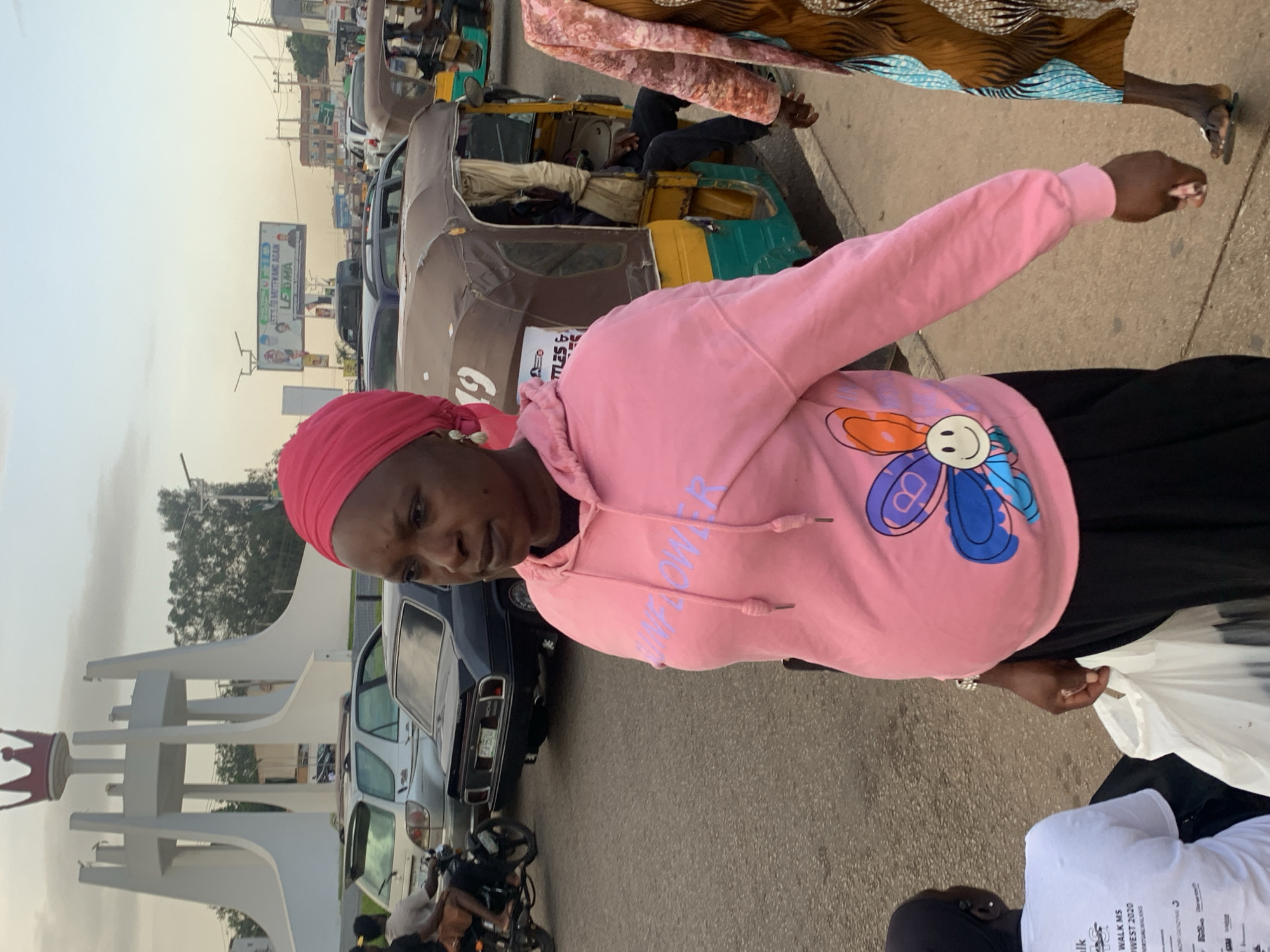In Jos, Nigeria, a woman wearing a pink hooded sweatshirt with a sunflower graphic and a pink headscarf stands on a street. She carries a white plastic bag. Behind her, a tricycle, a black car, and several other vehicles are visible, indicating traffic. In the background, a white monument topped with a red crown stands prominently, and a billboard displaying political figures is mounted above the road. The sky is overcast with a light haze.