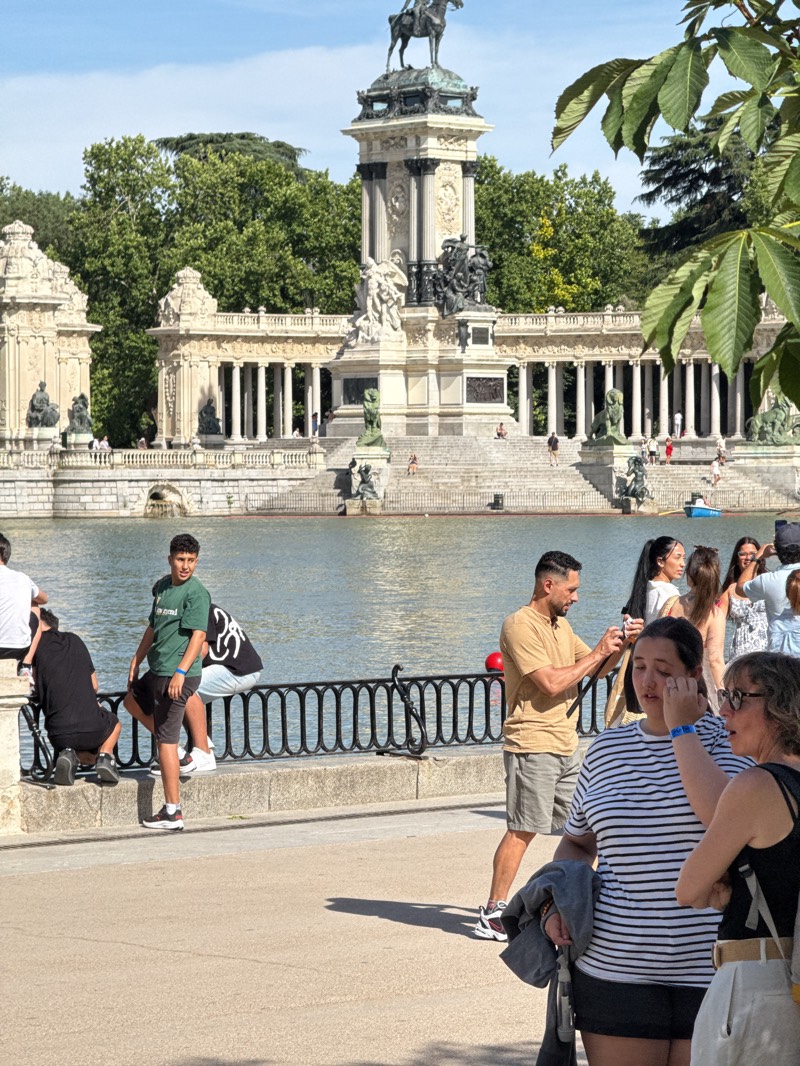 This outdoor image captures a lively scene at the Monument to Alfonso XII and the Grand Pond in El Retiro Park, Madrid, Spain, under bright daylight.
The central focus is the grand, neoclassical Monument to Alfonso XII, situated at the far end of a large, placid pond. The monument features a towering pedestal topped with an equestrian statue of Alfonso XII. Below, a colonnaded semi-circular structure with numerous classical sculptures and statues extends around the base, creating a majestic backdrop. Wide steps lead from the monument down to the water's edge, where several people are visible, some sitting on the steps, others walking. A few small boats can be seen on the pond, suggesting recreational activity.
In the foreground, a paved walkway runs parallel to the pond, separated by a black metal fence. A diverse group of people occupies this area. On the left, several individuals are relaxing near the fence, some sitting on the stone edge, others kneeling. A young man in a green t-shirt with partially visible white text and shorts stands near the fence, looking slightly left. His black and white sneakers are visible. Further right, a man in a tan t-shirt and shorts stands holding a phone, seemingly capturing a photo or video. Next to him, a woman in a striped t-shirt holds a jacket while talking to an older woman in a black top and glasses. Behind this group, more people are gathered, some facing the monument, others interacting amongst themselves. A red ball rests on the fence near the man with the phone.
The scene is illuminated by strong sunlight, casting distinct shadows on the walkway. Lush green trees frame the monument and fill the background, contributing to the park-like atmosphere. The weather appears warm and sunny, indicated by the attire of the people and the bright lighting.