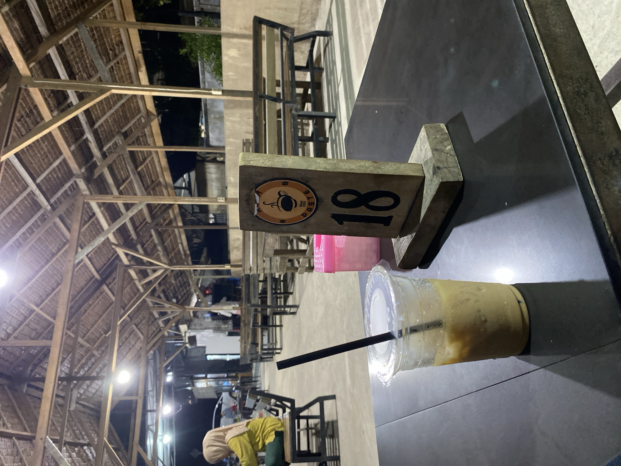 The image depicts an outdoor restaurant setting in Gampongaceh, Indonesia, at night. The roof structure is made of wood beams and thatch. Numerous metal tables and chairs are arranged in rows. In the foreground, a black metal table holds a plastic cup with a straw, containing a yellow beverage. A wooden table marker with the number "18" and a logo is also present. In the background, a person wearing a yellow top and hijab is near a table. The overall atmosphere is illuminated by artificial lights.