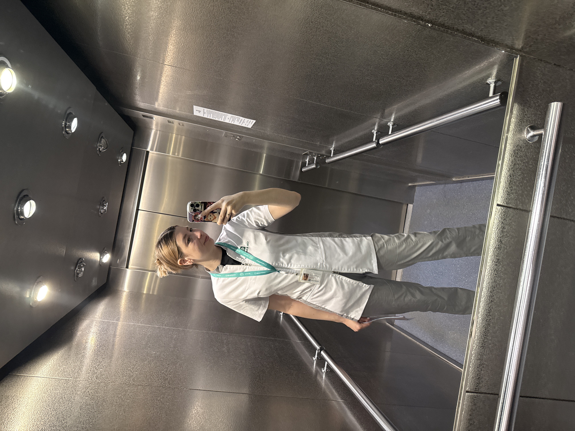 A person stands in a metal elevator. The person, wearing a white lab coat, light grey pants, and a lanyard around their neck, holds a phone up as if taking a selfie. The elevator interior is silver-colored, with metal walls and a ceiling featuring recessed lighting. A horizontal handrail is visible along one wall, and there is a metal panel running along the top of the walls. The location is not identifiable based on the image.