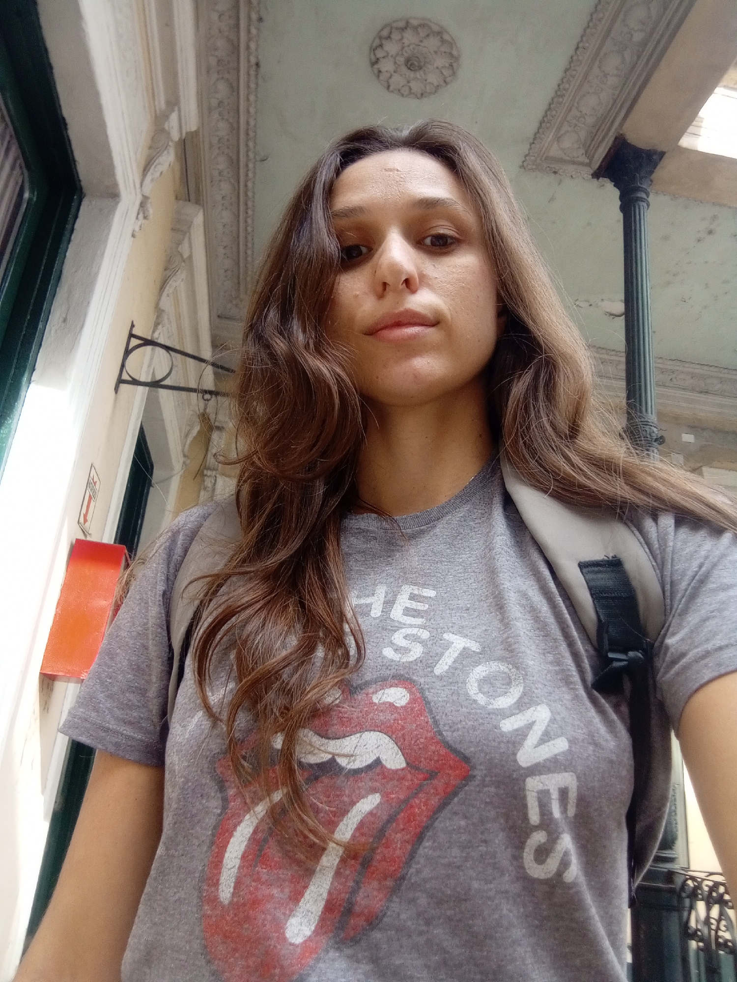 A woman wearing a grey Rolling Stones t-shirt and a backpack is standing underneath a covered walkway. The walkway has a decorative ceiling and columns. A building with a window and a red sign is visible on the left. The exact location in Buenos Aires, Argentina cannot be determined from the image alone.