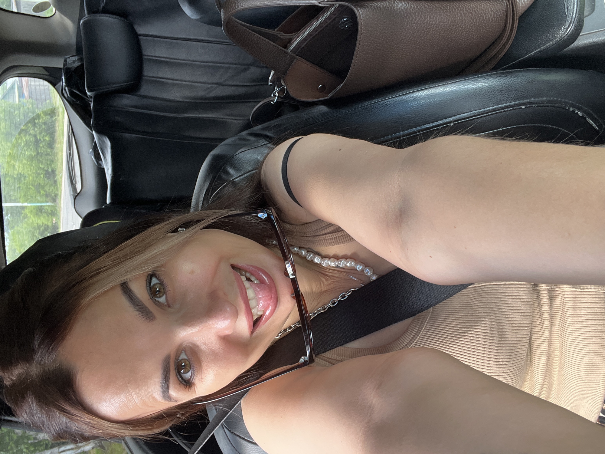 A woman is taking a selfie in the passenger seat of a car. She has brown hair with blonde highlights and is smiling with her tongue out. She is wearing a beige ribbed tank top, a pearl necklace, and silver chain. She is holding a pair of glasses in her right hand. A brown leather purse is visible on the seat next to her. The interior of the car, including black leather seats and a seatbelt, is also visible. The background shows greenery and part of a building through the car window.