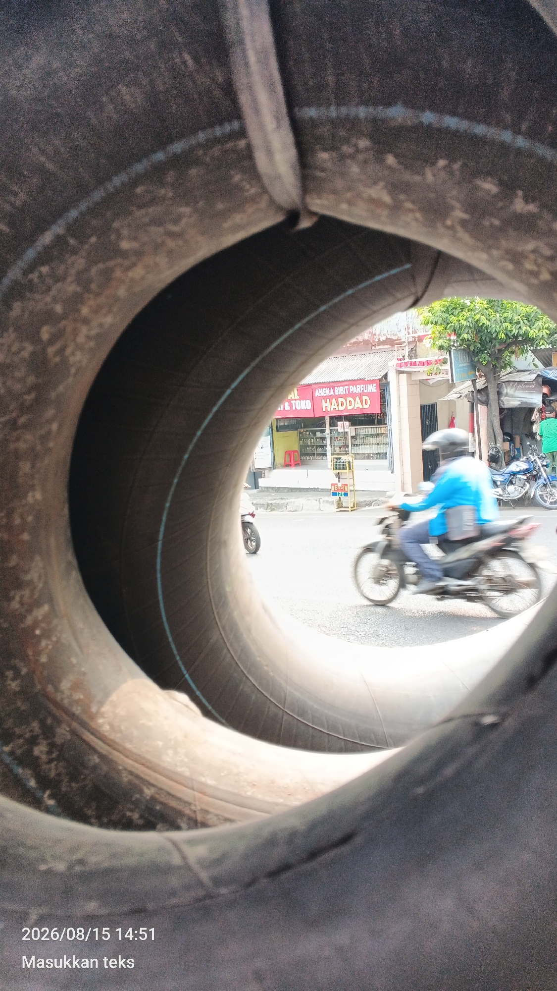 The image, taken on August 15, 2026, at 14:51, is a close-up view through the center of a large, dark-colored tire. The foreground is dominated by the textured, rubbery surface of the tire's inner wall. Through the opening, a street scene is visible in the mid-ground and background. A person wearing a blue jacket and a black helmet is riding a motorcycle in the mid-ground, moving from left to right. The motorcycle is slightly blurred, indicating motion. To the left of the motorcycle, another motorcycle is partially visible. In the background, there are buildings with signs, including one that reads "ANEKA BIBIT PARFUME" and "HADDAD." Trees with green foliage are also visible behind the buildings. The scene appears to be in an urban environment, likely a street in Malang, Indonesia, suggested by the presence of a local business sign. The ground is paved, and the overall lighting suggests it is daytime.