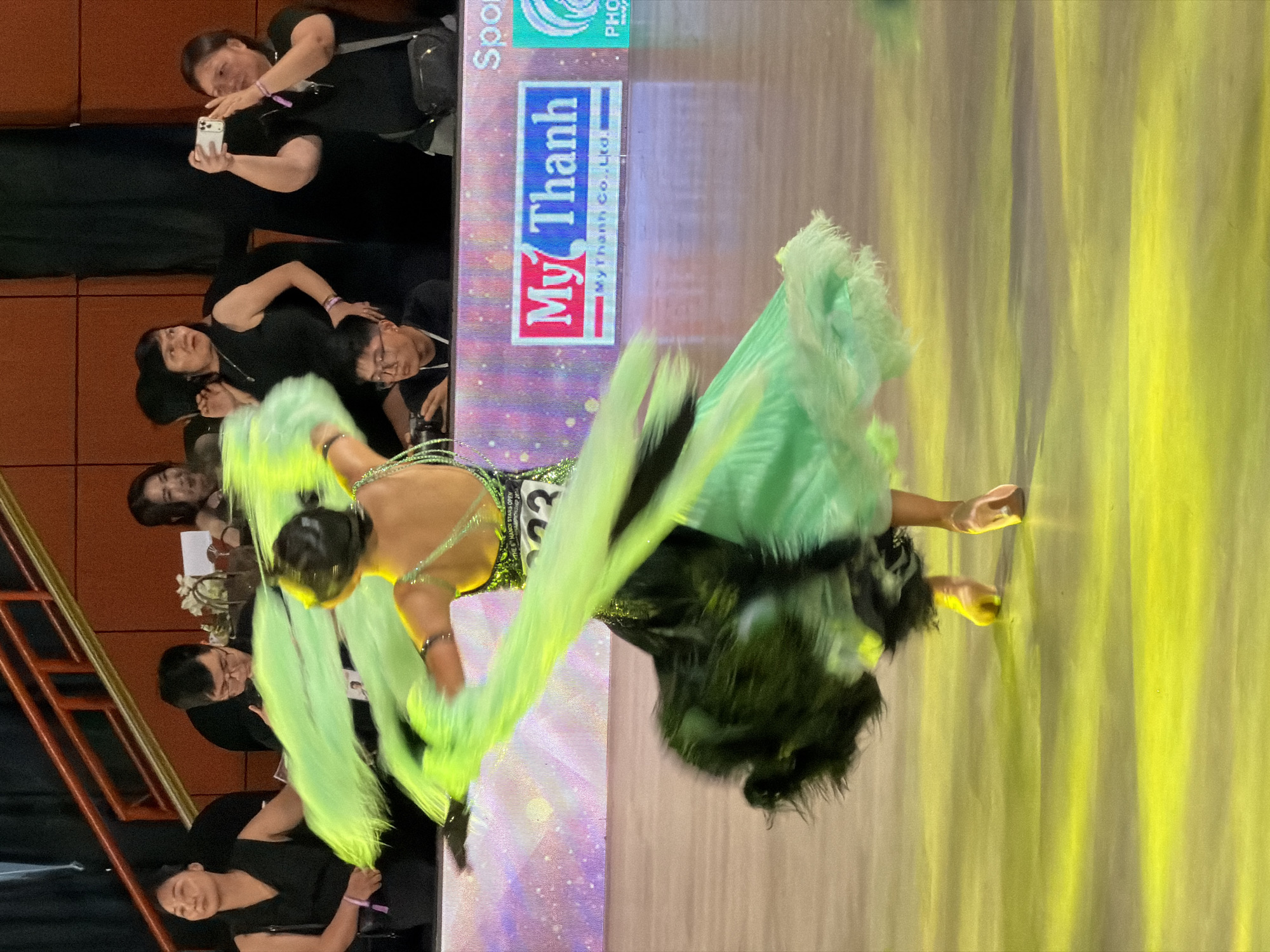 The image captures a moment during a ballroom dancing competition, likely held indoors at an event venue in Hà Nội, Vietnam. The central focus is a female dancer in mid-performance, her vibrant green and black sequined dress creating a blur of motion as she spins. The dress features flowing, feathery green accents that extend outwards with her movement. Her hair is pulled back, and she wears sparkling jewelry on her arms. The dancer's feet, clad in light-colored dance shoes, are visible as she balances on the wooden floor, which is illuminated by a strong yellow light, creating a vibrant, almost ethereal atmosphere.
In the background, spectators are seated behind a barrier, observing the competition. Among them, a woman in a black sleeveless top holds a smartphone, actively taking a photograph of the dancer. To her left, another woman in a black top looks towards the stage with a surprised expression, while a man with glasses is seated closer to the stage, also observing. Further to the left, a woman in a black sleeveless top sits with her hand resting on the barrier, watching the performance. Behind the spectators, a wooden railing and a red carpeted staircase are visible, suggesting the seating arrangement is tiered. A digital billboard is mounted behind the dancer, displaying the "My Thanh" logo, possibly indicating a sponsor of the event. The overall scene depicts a lively and dynamic atmosphere characteristic of a dance competition.