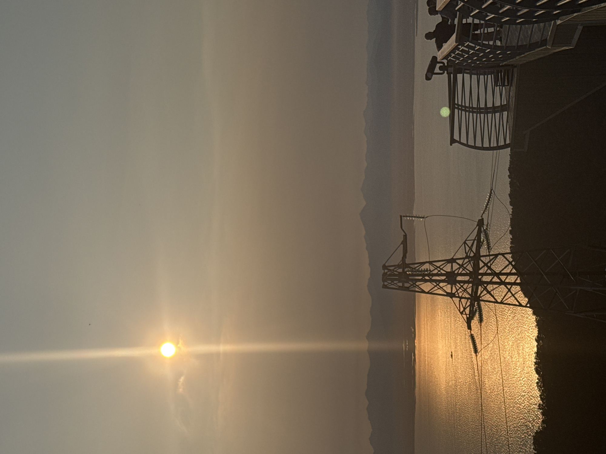 An outdoor scenic view shows a body of water reflecting the setting sun. A power line tower stands in the foreground, silhouetted against the water and sky. In the middle ground, a small boat is visible on the water. In the background, a mountain range stretches across the horizon. On a viewing platform on the right side of the image, two people are standing, looking out at the view, with one person appearing to look through a telescope. The sky is overcast, with a bright sun partially obscured by clouds, creating a strong vertical light beam. The scene appears to be located in Petropavlovsk-Kamchatsky, Russia, known for its dramatic natural landscapes and proximity to the Pacific Ocean.