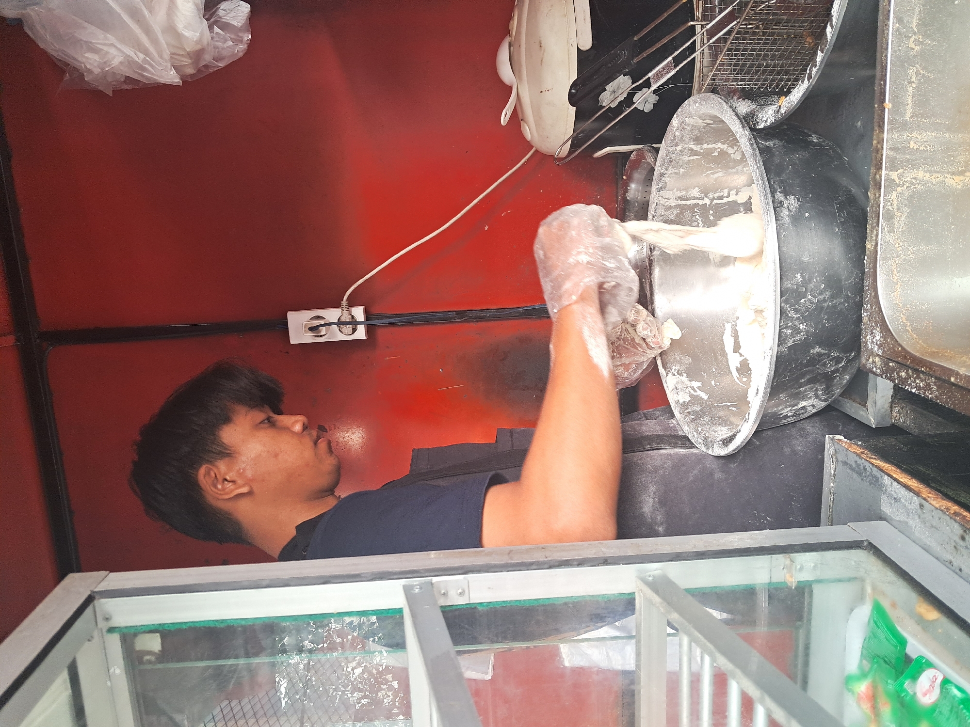 A young man, wearing a dark blue t-shirt and a dark apron, stands in what appears to be a food stall. He is in the process of preparing food, with his gloved hands immersed in a large metal bowl filled with a white, dough-like substance. The bowl is placed on a metal counter. To the right of the bowl, a deep fryer basket is visible, suggesting the food is likely to be fried. Behind him, the wall of the stall is painted a vibrant red. An electrical outlet with a white cord plugged into it is mounted on the red wall. On the far right, a white appliance, possibly a rice cooker, is present. To the left, a glass display case with metal framing contains some green packets of what appear to be condiments. The overall impression is of a busy, functional workspace within a small food vendor setup, possibly in Klaten, Indonesia, a city known for its diverse culinary offerings.