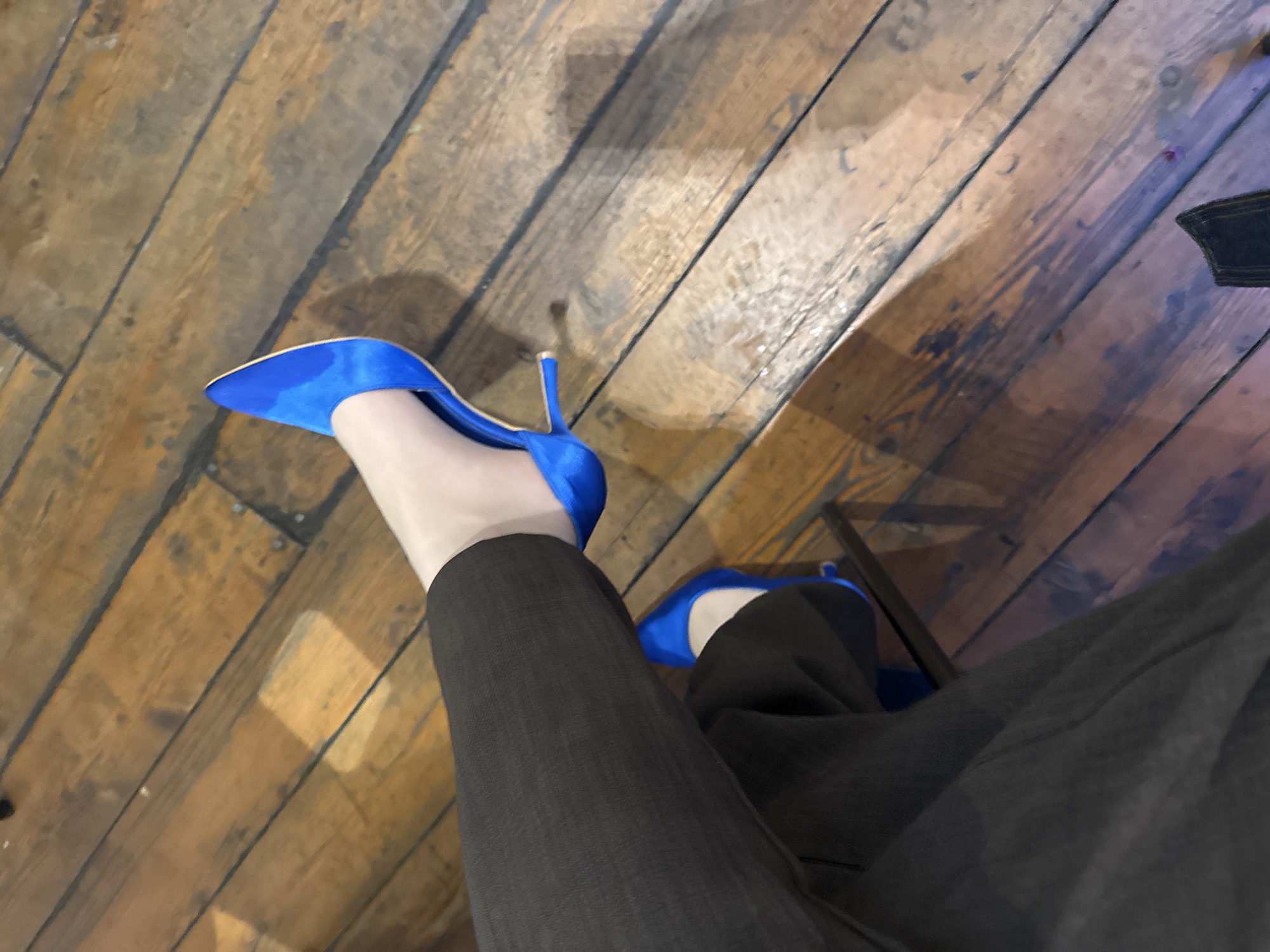 A downward-facing, medium-shot photograph depicts a person wearing dark, likely charcoal gray, slacks and vibrant blue stiletto heels on a weathered wooden floor. The floorboards show signs of wear and variations in color, suggesting age and use. The pants cover the legs, ending at the ankles where the blue heels are displayed. The background is a blur, but a dark object in the upper right corner suggests a table or a piece of furniture. The lighting is soft and diffused, giving a warm hue to the wooden floor. The focus is sharp on the shoes and pants, suggesting they are the primary subjects of the image. There is no indication of the location being Yerevan, Armenia or any other specific place. No actions or interactions are discernible.