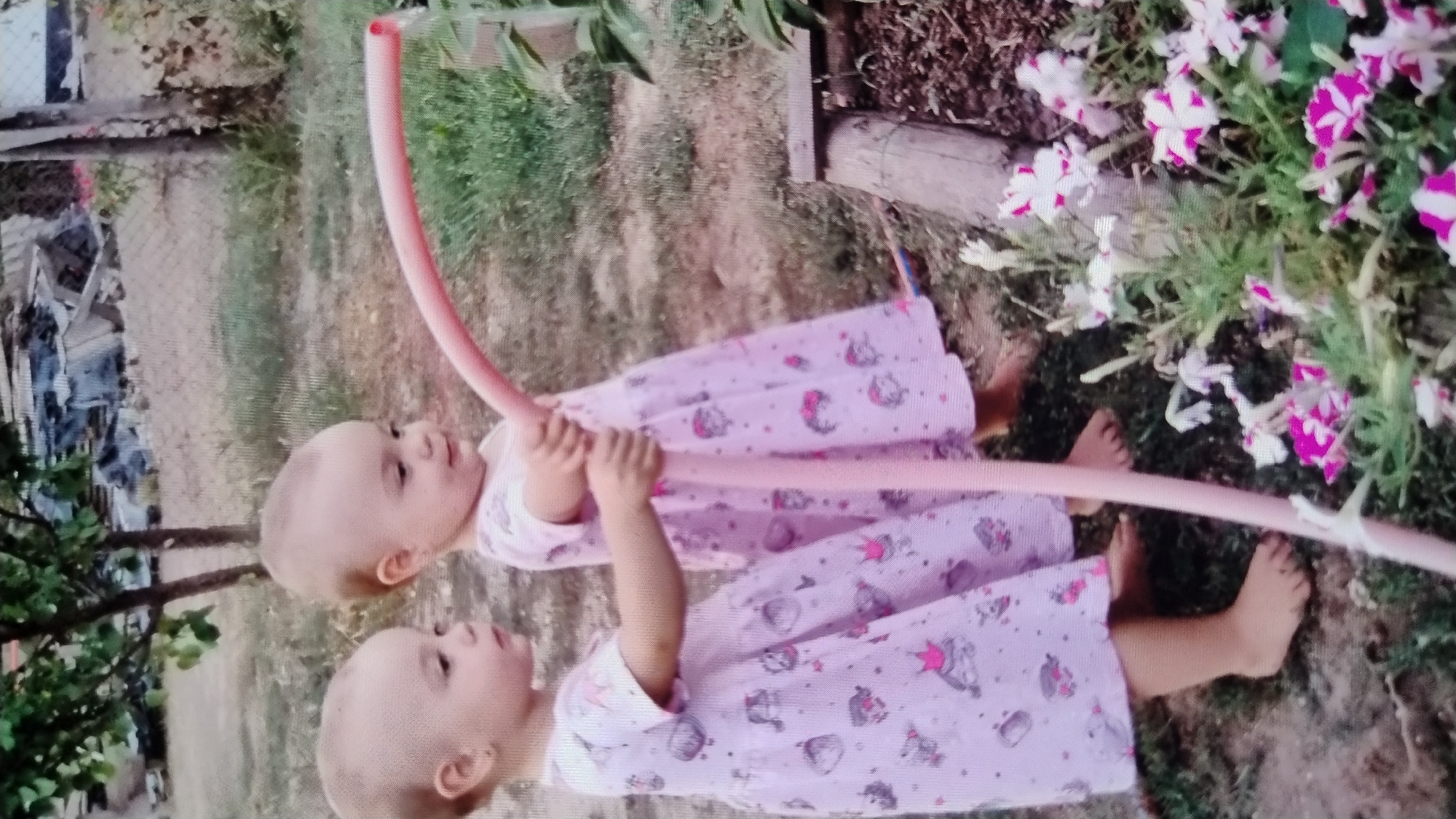 Two young Caucasian female toddlers, each wearing a light pink dress with a repeating pattern of cartoon characters and crowns, are standing next to each other outdoors. They are both holding a pink garden hose, with water spraying from the end. The child on the right is looking towards the hose and smiling, while the child on the left is looking to the left of the frame. They are both barefoot. The environment appears to be a backyard with dirt ground, grass, and some plants and flowers. There are also some trees and a fence in the background, along with debris that suggests a possibly neglected or damaged area. The image does not provide any specific details that would allow for localization to Mykolaiv, Ukraine.