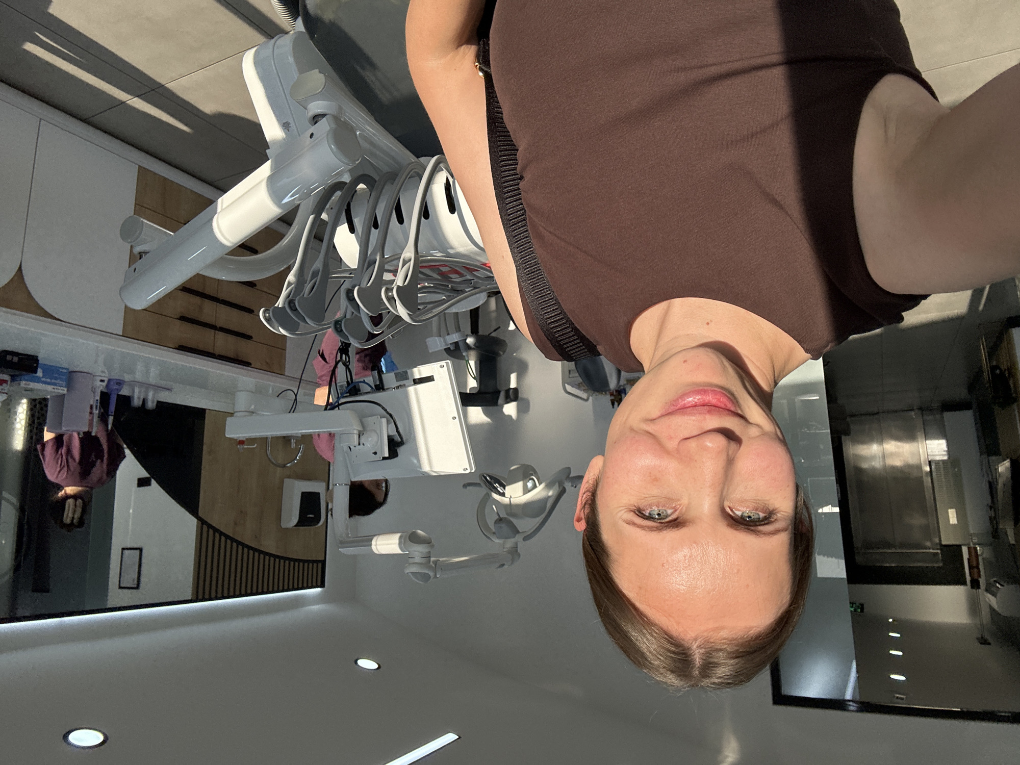 A selfie depicts a fair-skinned woman with light brown hair, wearing a brown tank top, in a dental clinic. The clinic appears modern with white walls, recessed lighting, and dental equipment. Reflections of a person and a hallway are visible in a large mirror. The location is presumed to be Fethiye, Turkey.