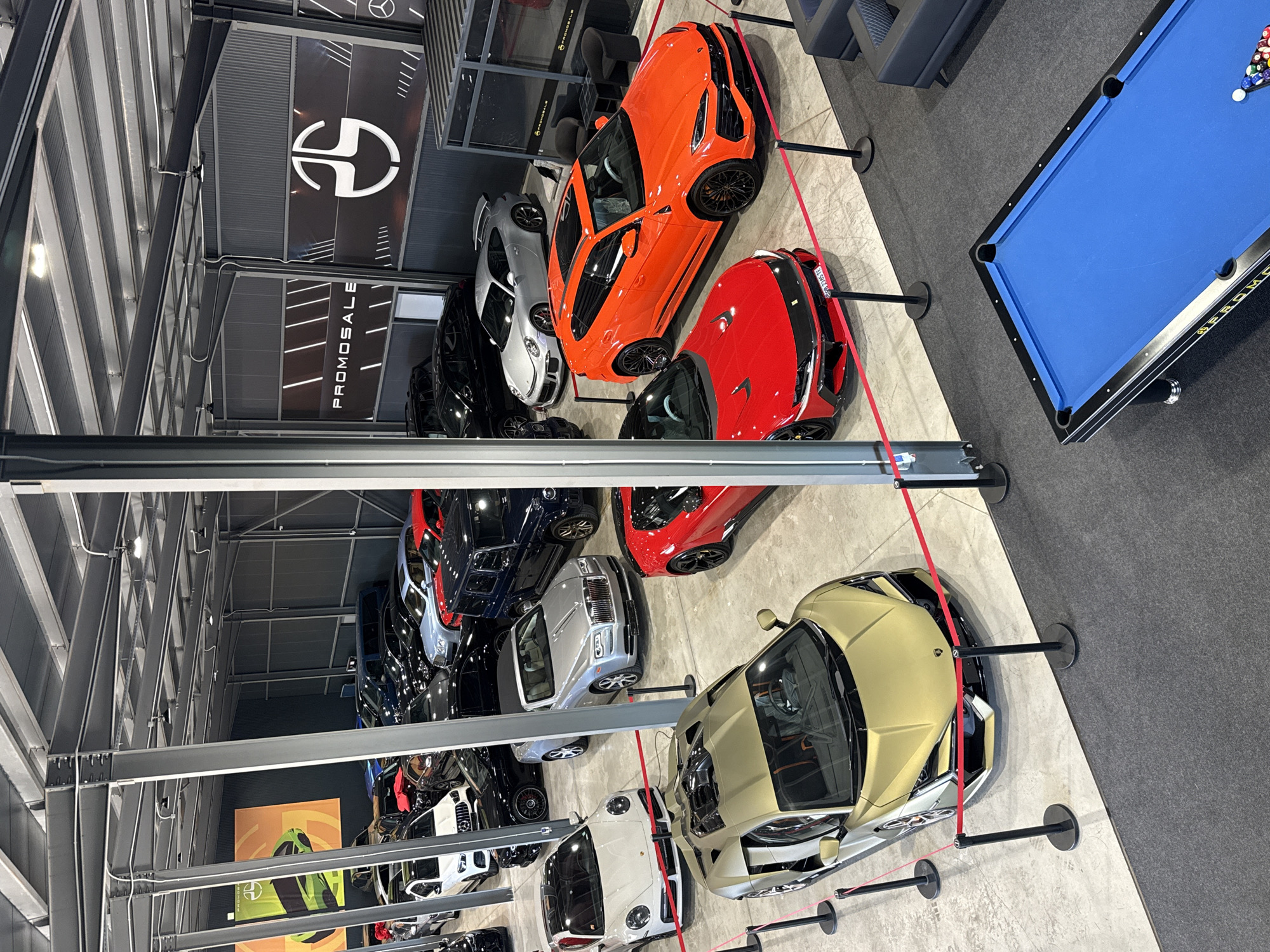 A high-angle shot shows the interior of a car dealership in Plovdiv, Bulgaria. A variety of luxury cars, including an orange SUV, a red sports car, a gold sports car, and several black and white vehicles, are displayed on a grey concrete floor. A stanchion with a red rope separates the cars from the rest of the room. A pool table with blue felt is located in the upper right corner of the image on a dark grey carpet. A company logo is visible on the upper wall. Steel beams and supports are present in the building's structure.