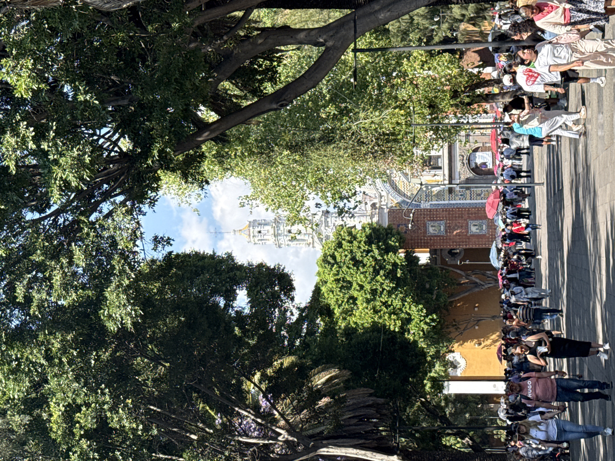The image shows a crowd of people walking along a street in Puebla, Mexico. The street is lined with trees, and in the distance, the towers of the Templo de San Francisco can be seen. The people are dressed in a variety of clothing styles. Many carry umbrellas.