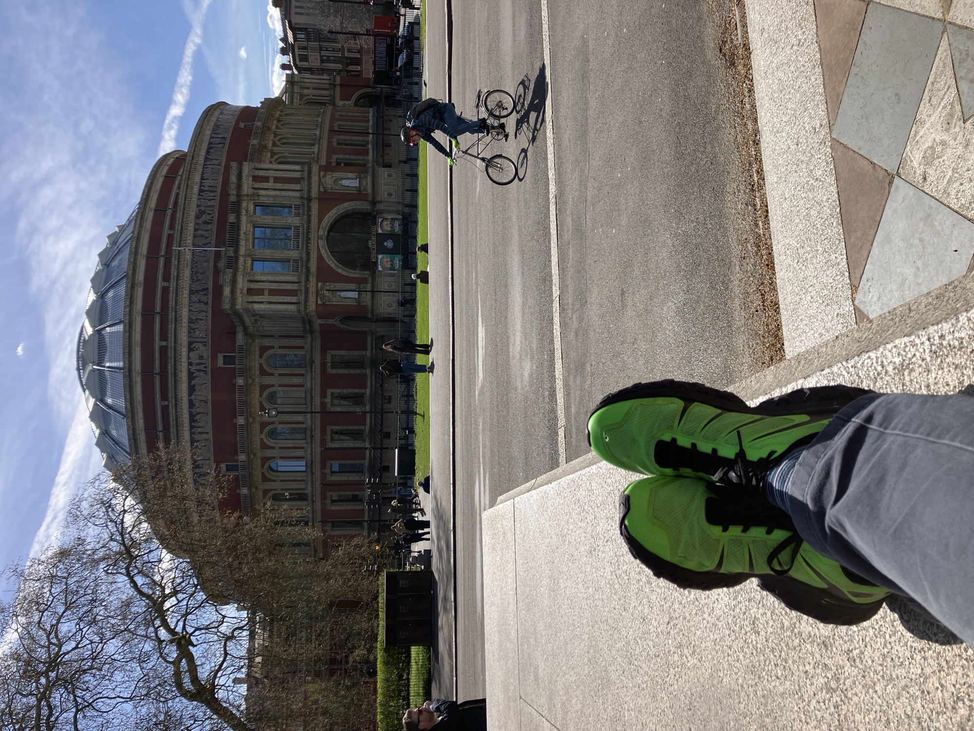 An outdoor urban scene in London, United Kingdom, specifically facing the Royal Albert Hall in Kensington. In the immediate foreground, the feet of an individual are visible, clad in bright neon green and black athletic shoes and dark grey trousers, resting on a light grey, textured stone surface.
A paved road, Kensington Gore, occupies the midground. A male cyclist, wearing a helmet, striped long-sleeved top, jeans, a backpack, and bright green gloves, is actively pedaling a small-wheeled bicycle from right to left across the road, casting a long shadow.
The background is dominated by the circular, red-brick facade of the Royal Albert Hall, featuring a prominent glass dome roof, multiple arched windows, and a decorative frieze depicting figures. A British flag is visible flying from the building. A large, bare-branched tree stands to the left, partially obscuring the building's edge. A grassy area lies between the road and the Royal Albert Hall, where several pedestrians are visible standing and walking. The sky is clear blue with sparse white clouds and a single contrail. The scene is illuminated by direct sunlight.