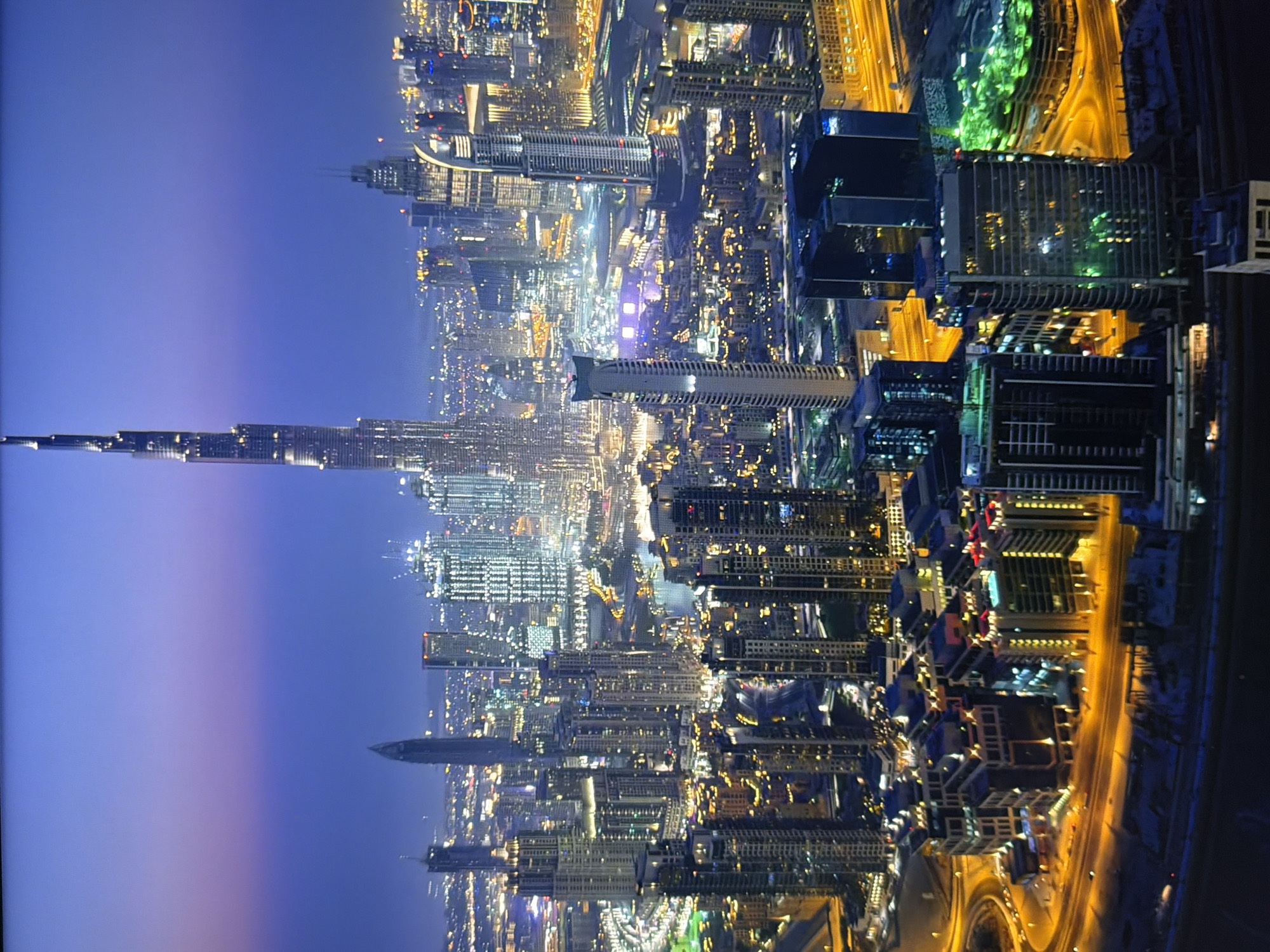 The image depicts a panoramic view of a modern cityscape at dusk. The sky transitions from a deep blue at the top to a lighter, purplish hue near the horizon. A towering skyscraper, likely the Burj Khalifa due to its distinctive spire, dominates the center of the frame. Surrounding it are numerous other skyscrapers of varying heights and architectural styles, many of which are illuminated with warm yellow and white lights. Roads with visible traffic, appearing as streaks of light, weave through the city, highlighting the urban infrastructure. In the foreground, several lower-rise buildings and complex intersections are visible, also lit up. The overall impression is one of a vibrant, densely populated metropolis at night. The question regarding the city of Kyiv, Ukraine, is not supported by the visual evidence in the image.