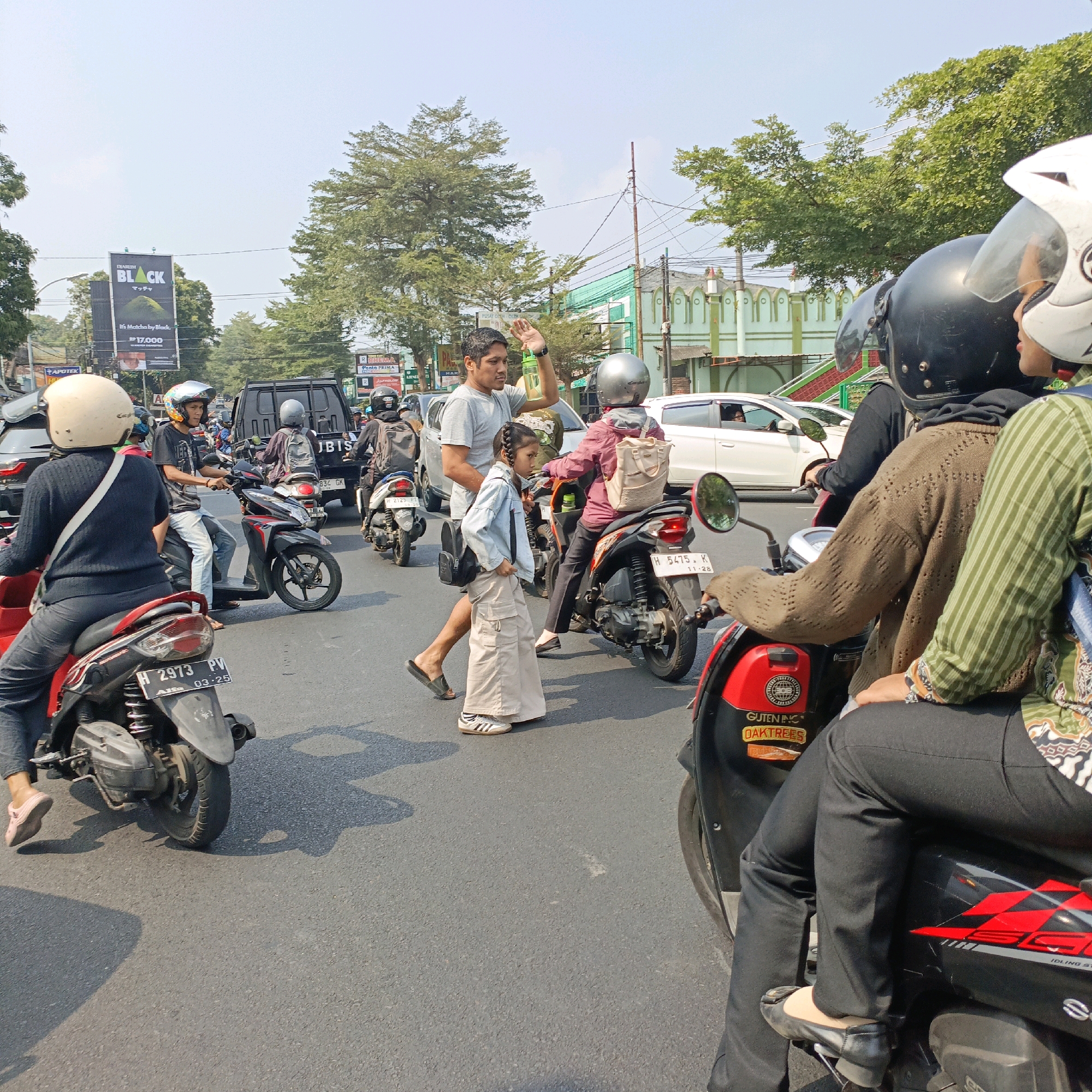 The image captures a street scene in Salatiga, Indonesia, characterized by moderate traffic congestion. The perspective is from a low angle, looking forward as if from the back of a motorcycle. Multiple motorcycles and a few cars are visible, navigating a paved road under a clear, bright sky.
In the foreground, the rear of a red and black motorcycle is prominent, with a person wearing black pants and black ballet flats seated on it. To the left, another motorcycle in red and black is partially visible, with a person wearing a black long-sleeved shirt and black pants, also on a motorcycle.
Moving towards the center, a man in a grey t-shirt is walking beside a young girl in a light blue denim jacket and a long, beige skirt. The man is waving, and the girl is looking ahead. They appear to be pedestrians in the midst of the traffic. Behind them, several other motorcycles are present, carrying riders with helmets.
Further back, a black SUV is stopped in traffic, and beyond that, more vehicles and motorcycles fill the roadway. The surroundings include trees with green foliage and buildings, one of which is light green with decorative white elements. A large black banner advertising "DIARIUM BLACK" and "Matcha by Black" is also visible on the left, indicating commercial activity. The overall atmosphere suggests a typical day on a busy street in an Indonesian city.