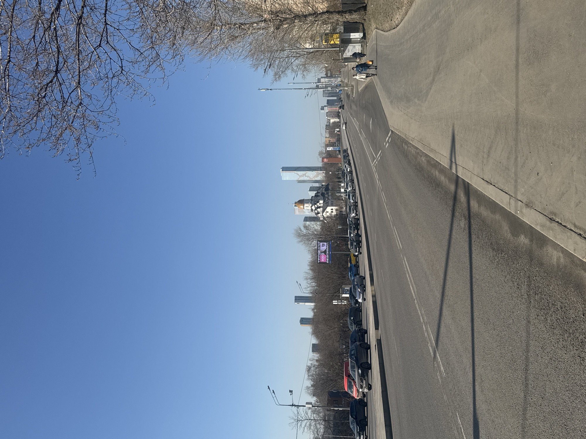 The image depicts a long, straight road in Moscow, Russia, heavily congested with stationary cars. Tall buildings and a church with gold domes can be seen in the distance, suggesting an urban setting. Bare trees line the roadside, and a clear blue sky is visible. Two pedestrians are walking along the right side of the road. A shadow from a street lamp is visible on the sidewalk.