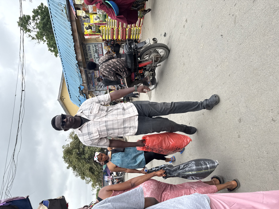 The image depicts a bustling street scene in Ajah, Nigeria, characterized by pedestrian and motorcycle traffic alongside market stalls.
In the foreground, a Black male, estimated to be between 25-35 years old, walks towards the left of the frame. He wears a light-colored plaid short-sleeve button-up shirt over a dark undershirt, dark jeans, and dark closed-toe sandals. He has dark sunglasses on and a short, dark haircut. He carries a large, full red plastic bag in his right hand and a smaller black plastic bag in his left hand. To his immediate left, partially obscured, are other pedestrians, including a female in a pink patterned garment carrying a black plastic bag and another in a light grey t-shirt.
In the midground, to the left of the central figure, a Black female, approximately 20-30 years old, walks away from the viewer. She wears a blue halter-neck top, dark leggings, and a white patterned headband, holding a small white water bottle. To the right of the central figure, a Black male, approximately 20-30 years old, sits on a red motorcycle, facing away from the viewer. He wears a dark patterned long-sleeved shirt.
The background features market structures. A building with a light blue corrugated metal roof runs along the right side of the street. Underneath, signs are visible, one advertising "JAMIE COLLEGE & CULTURAL ORGANIZATION" and educational degrees (B.Ed, B.A, Ed. Local & Int'l) with a phone number (08064974999). Another sign advertises "QUALITALL GOLD" with images of animals, positioned above stacks of yellow and green bags (likely feed or produce). A person in a red robe carrying a light blue basin is visible on the far right. Further left in the background, trees and more distant market activity are present. The ground is a light-colored concrete or paved surface, showing some wear. Overhead, power lines crisscross against a cloudy, overcast sky.