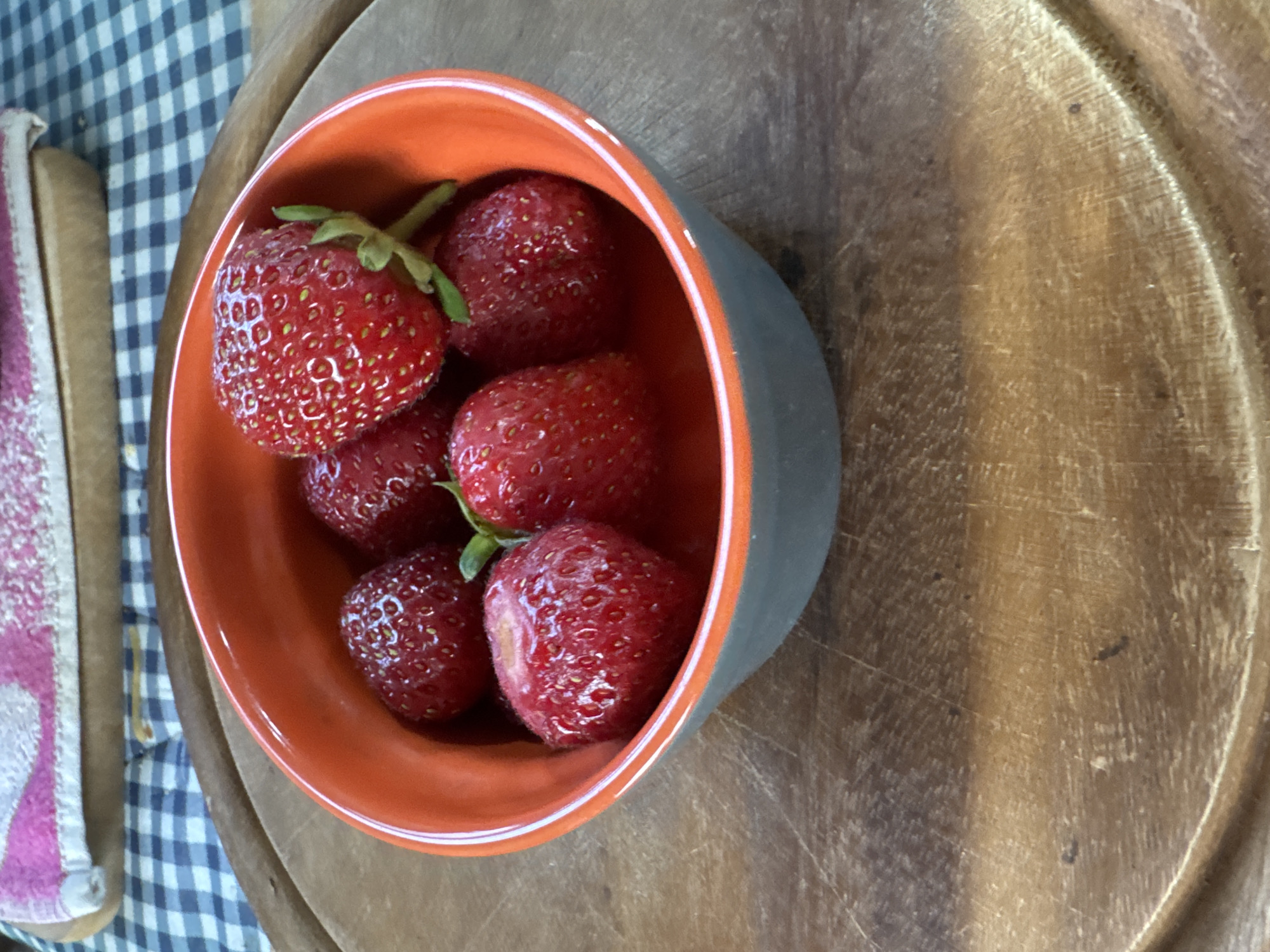A close-up shot shows a ceramic bowl filled with fresh strawberries. The bowl is predominantly orange on the inside and gray on the outside, with a white rim. It rests on a round wooden cutting board. In the background, a blue and white checkered cloth is visible, topped with a folded textile featuring pink and white patterns. The image appears to be taken indoors, possibly in a kitchen or dining area. There are no people or animals present in the scene, and no specific location in Odesa, Ukraine can be identified from the visual cues.