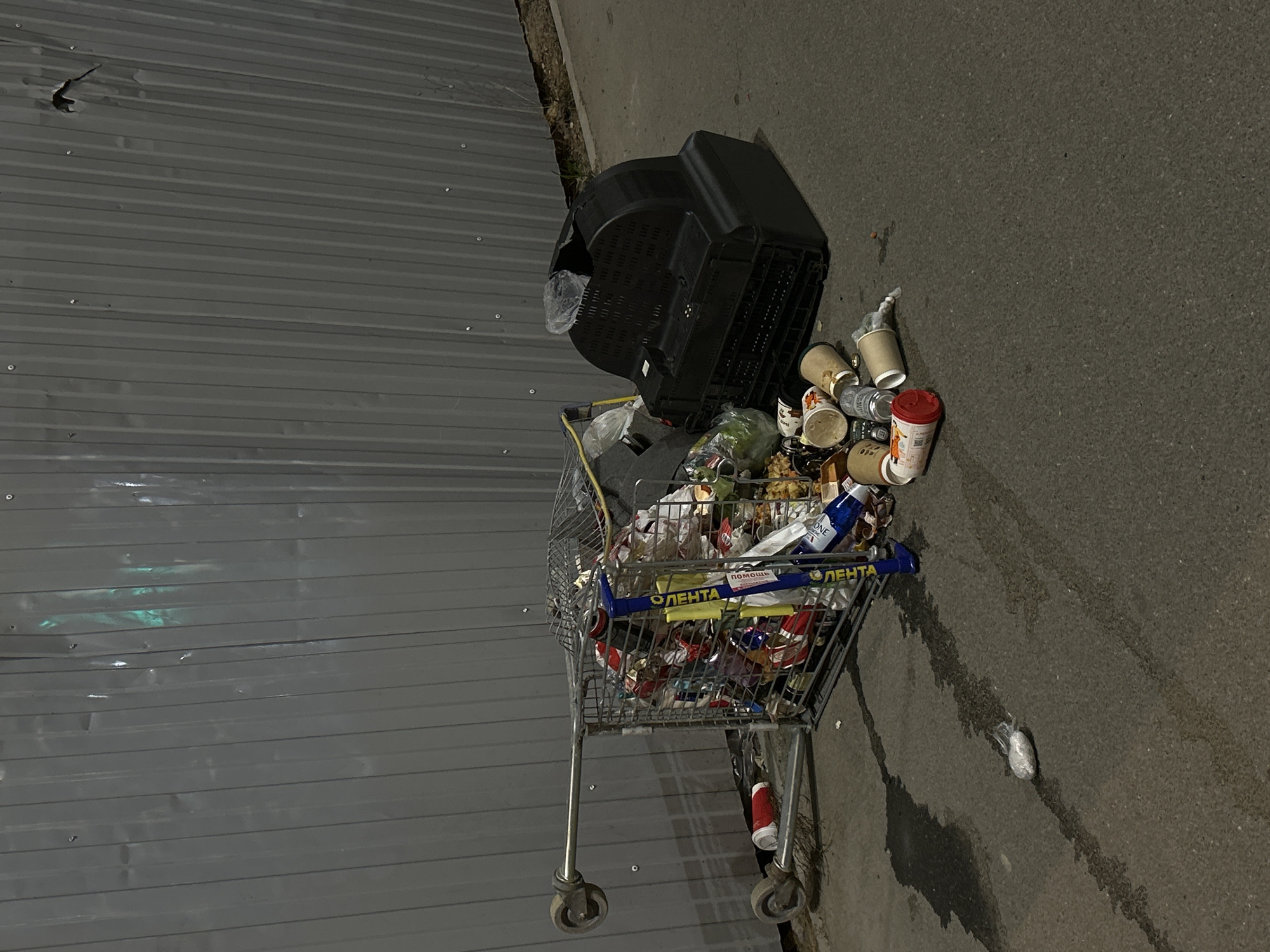 This nighttime photograph captures a scene of urban neglect in what appears to be Saint Petersburg, Russia. The main subject is a metal shopping cart, overloaded with various discarded items and refuse, positioned on a paved surface next to a dark, corrugated metal fence or wall.
An old, black cathode-ray tube (CRT) television set rests precariously on or beside the overflowing shopping cart, adding to the pile of garbage. The cart itself is filled with an assortment of trash including plastic bags, food packaging, bottles, and general debris, indicating a large amount of dumped waste. The handles of the shopping cart clearly display the Russian word "ЛЕНТА" (LENTA), identifying it as belonging to a prominent Russian hypermarket chain, and another label reads "ПОМОЩЬ" (POMOSHCH), meaning "help."
Scattered on the asphalt immediately around the cart and television are numerous disposable coffee cups of different sizes and colors, some with visible logos, along with other small pieces of litter. The paved ground also exhibits dark, wet stains or patches. The corrugated metal wall on the left side of the frame shows some minor dents or irregularities. The dim, artificial lighting suggests it is night, casting soft shadows across the scene. There are no people visible, and the overall impression is one of abandonment and urban decay.