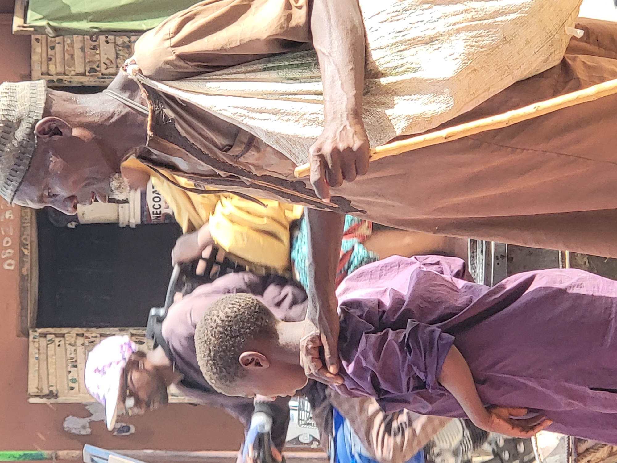 In Maiburiji, Nigeria, an elderly man wearing a knitted cap and a brown robe, carrying a bag and a wooden stick, stands with a child in a purple robe. Another person, wearing a white cap, stands nearby. A sign with the partial word "NECOA" is visible in the background.