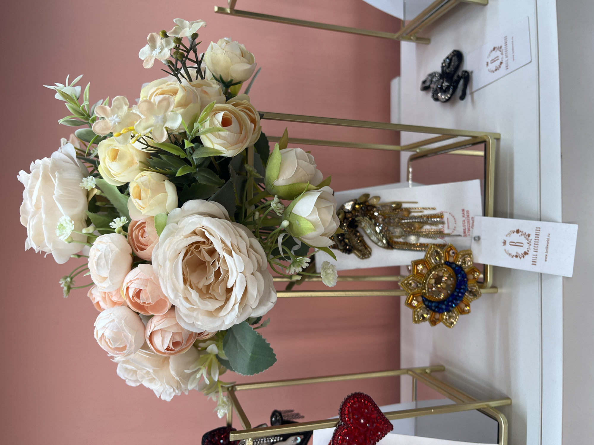 An indoor retail display features a bouquet of artificial flowers in pastel shades of cream, blush pink, and pale yellow, arranged in a white vase that sits on a white shelf. The vase is placed on a gold metal stand. To the left of the flowers, a gold metal display rack holds a red heart-shaped brooch adorned with rhinestones. To the right of the flowers, a similar gold metal stand showcases a brooch resembling a horse's head with dangling embellishments. Below the flowers, also on the shelf, are two more brooches: one circular with blue and gold beads and a crescent moon design, and another shaped like a black snake. The background is a solid pink wall. The overall impression is that of a small boutique or shop selling accessories. The specific location within the city of Svetly, Russia, is not discernible from the image itself.