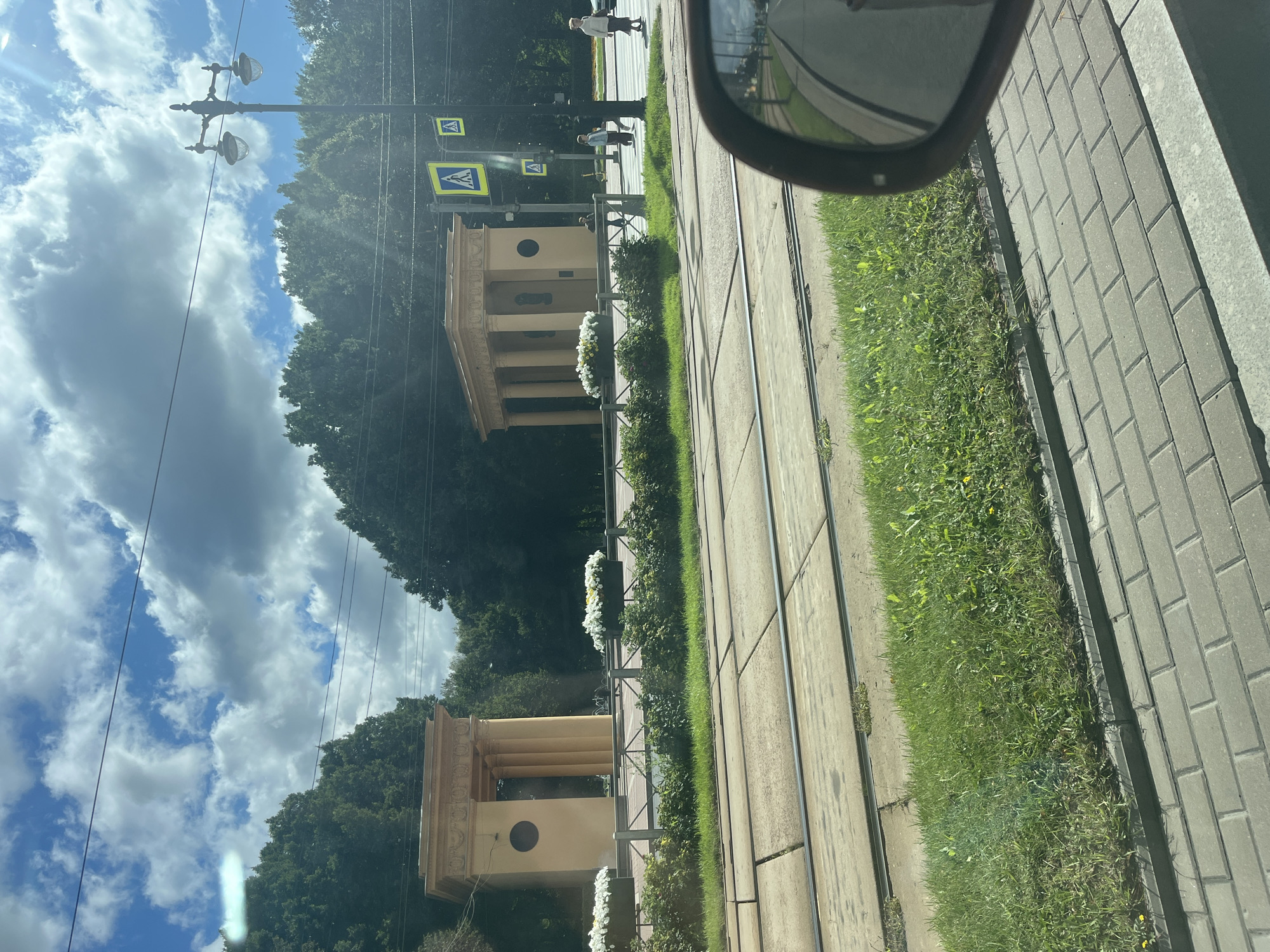 The image shows a sunny day in Saint Petersburg, Russia, with a view of two identical neoclassical pavilions with columns, likely part of an architectural ensemble. The pavilions are yellow, with a circular indentation on the front facade of each. They are situated on a grassy area bordered by a metal fence, with planters filled with white flowers in front of them. In the foreground, tram tracks are visible on a paved surface, with a strip of grass between the tracks and a sidewalk made of grey interlocking pavers. A lamppost with two lights stands to the right of the pavilions, next to pedestrian crossing signs. Several people are walking on the sidewalk in the background, and a car's side mirror is reflected in the lower right corner of the image. The sky is blue with fluffy white clouds. The overall impression is one of a well-maintained urban park or public space in Saint Petersburg.
